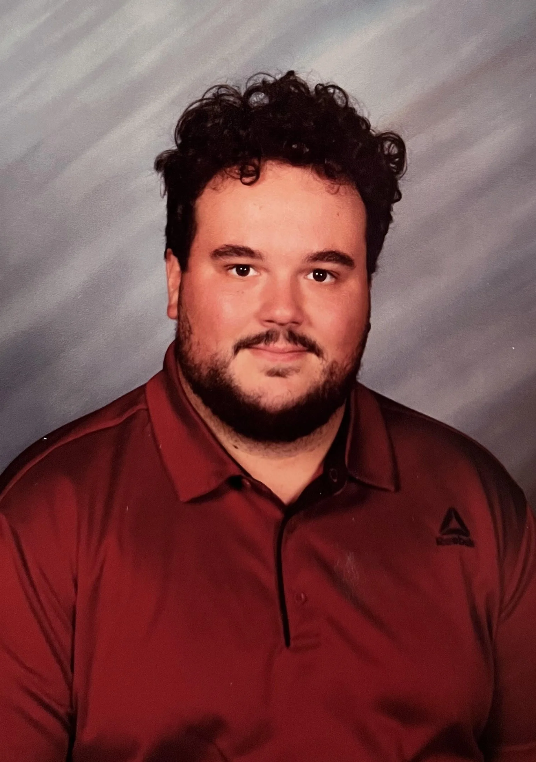A young man with curly dark hair, beard, and light skin, wearing a red Reebok polo shirt, poses for a portrait against a gray gradient background.