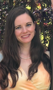 A woman with long brown hair and blue eyes smiling outdoors, wearing a pearl necklace and a sleeveless yellow dress, with greenery and flowers in the background.