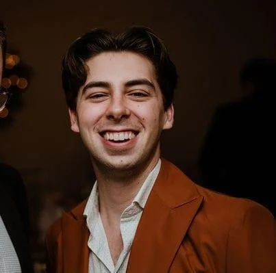 Young man with dark hair smiling, wearing a brown blazer and a light-colored shirt, at an indoor social event.