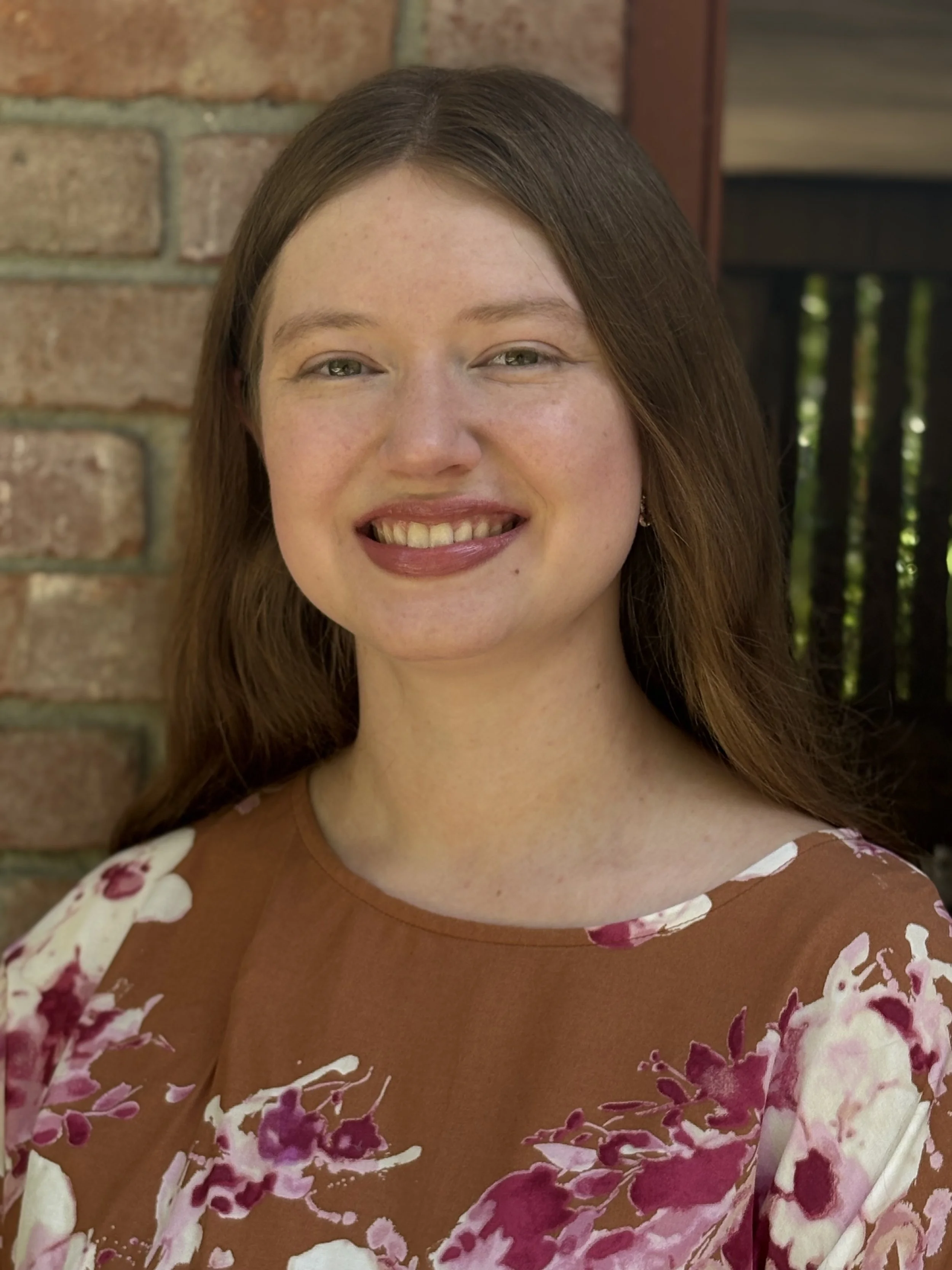 A woman with long, reddish-brown hair smiling outdoors against a brick wall and wooden fence, wearing a brown dress with pink and white floral patterns.