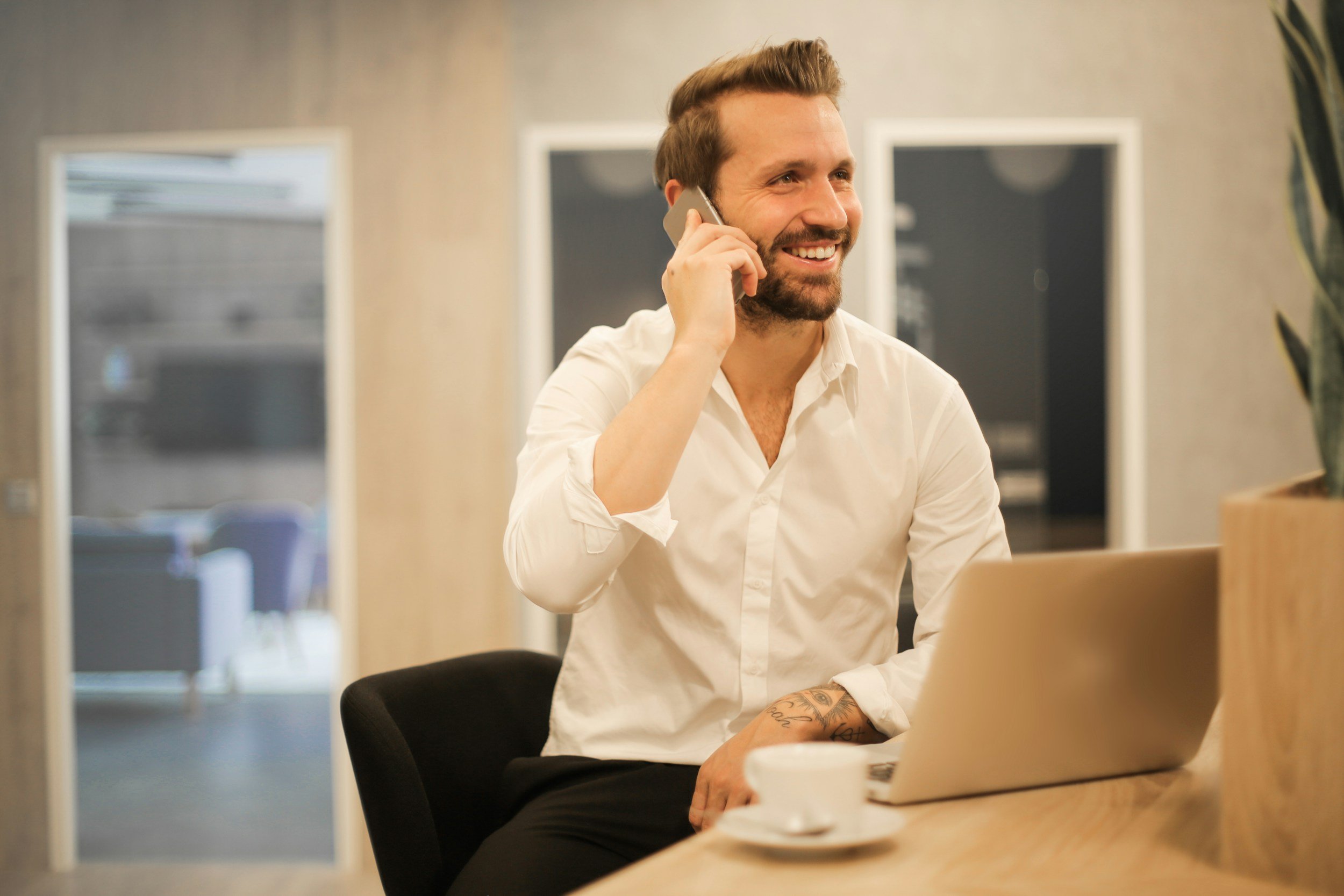 A man with brown hair and a beard, wearing a white shirt, is smiling and talking on a cell phone. He has tattoos on his arm and is sitting at a table with a laptop in front of him, in an indoor setting that appears to be an office or a cafe.