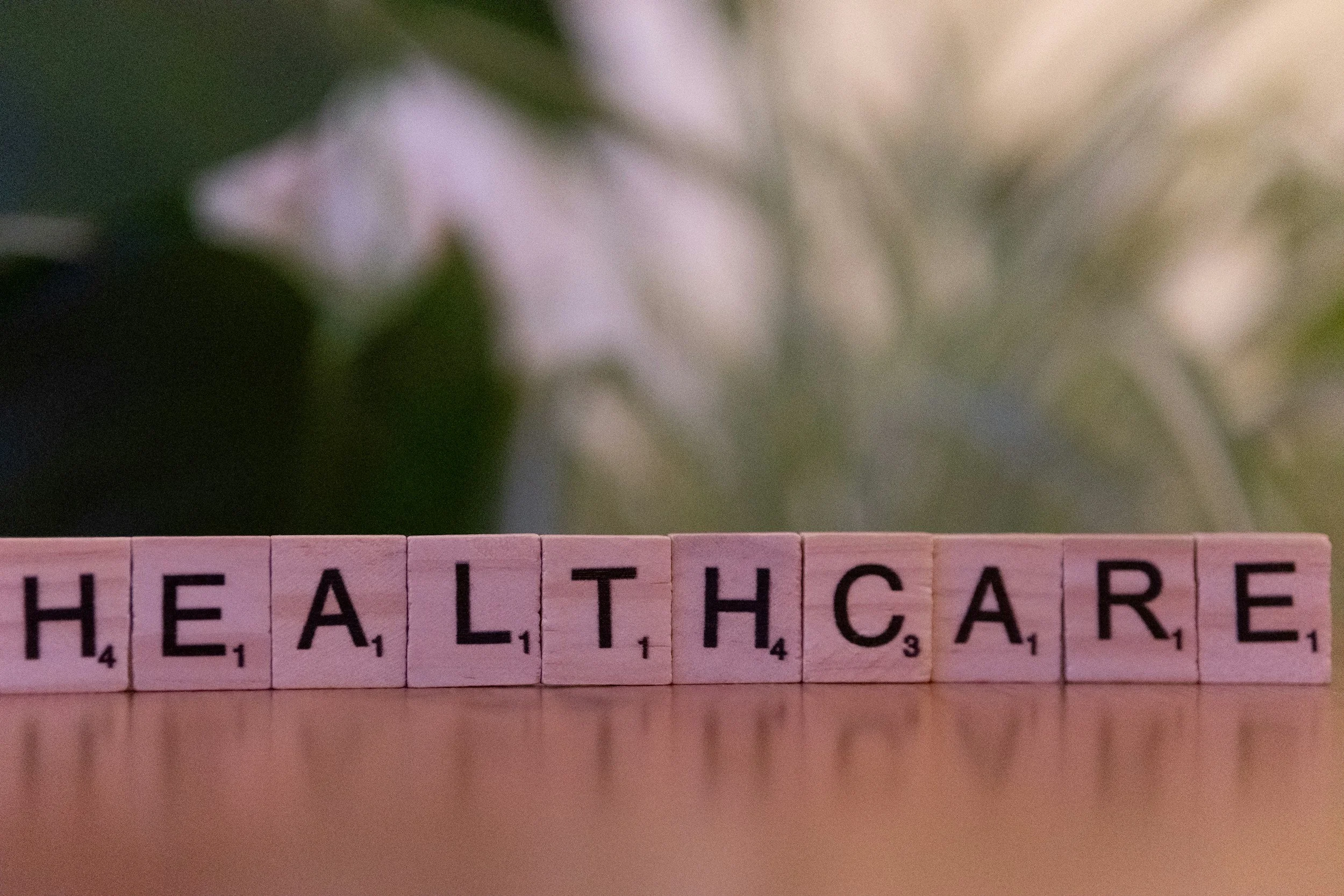 Pink letter tiles spelling out 'THE HEALTHCARE' with a blurred background of white flowers and green leaves.