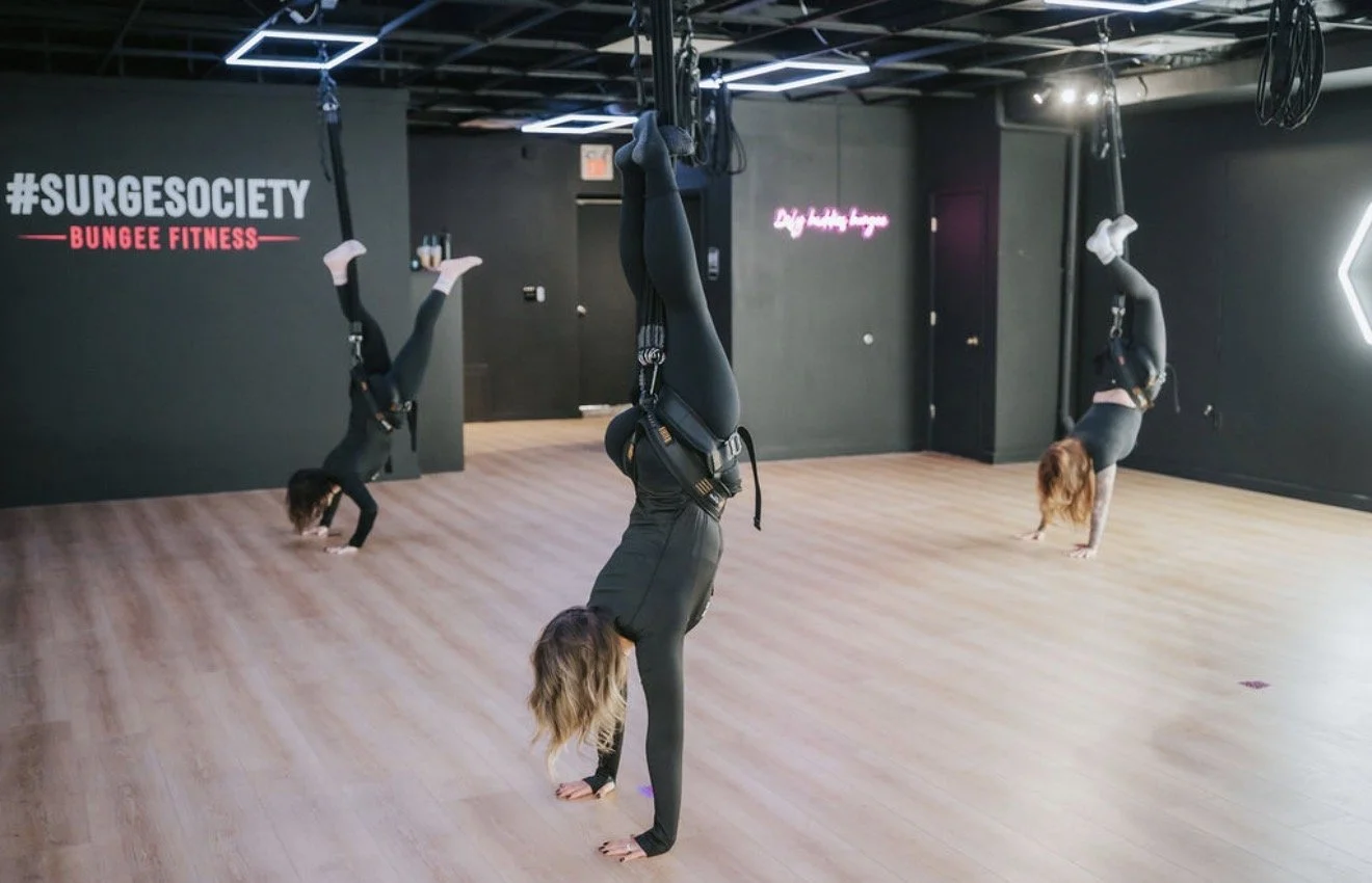 Three women practicing aerial yoga in a fitness studio with black walls, wooden flooring, and neon signs.