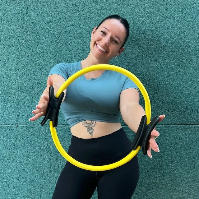 Young woman smiling and holding a yellow fitness hoop in front of a green wall.