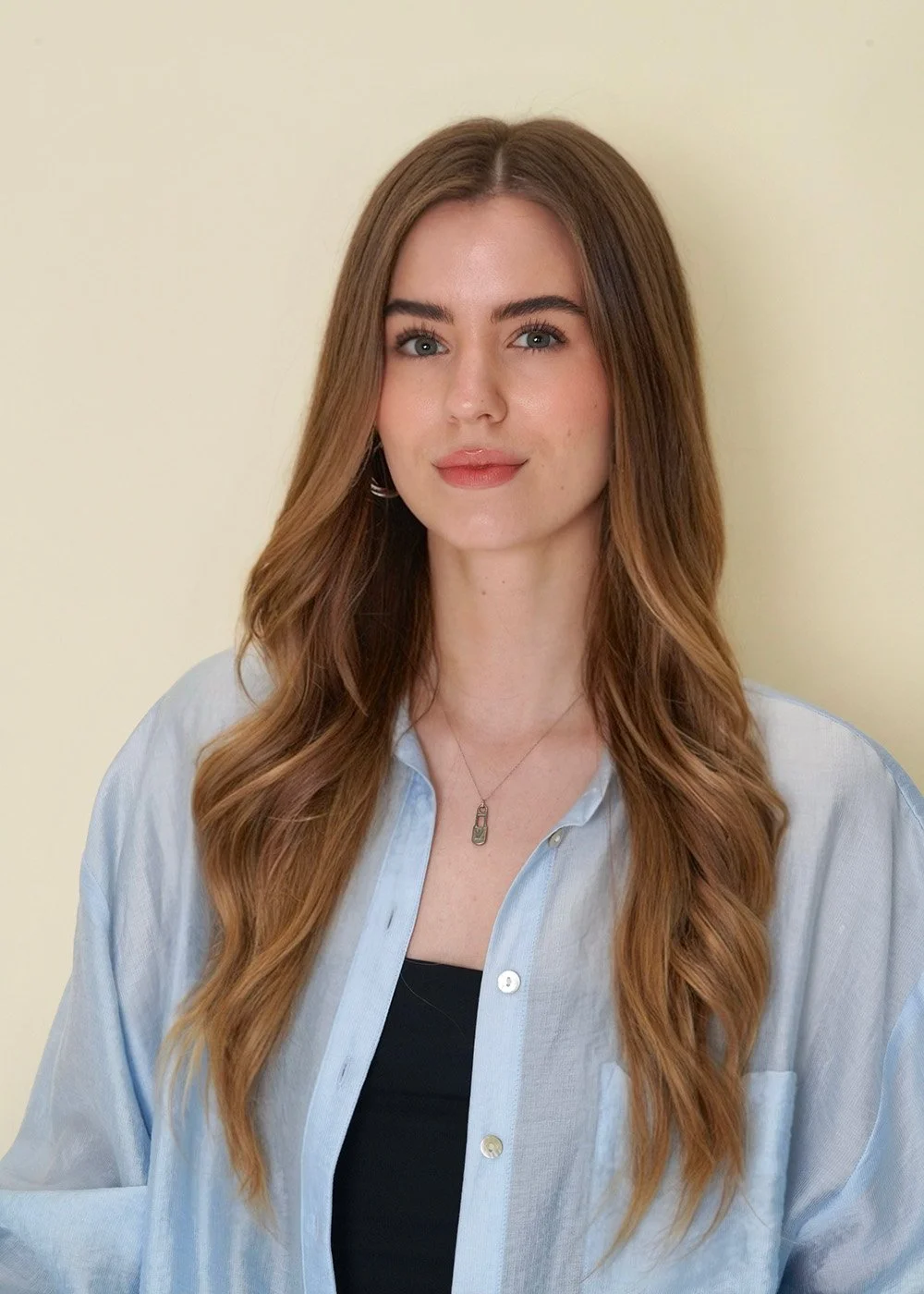 A young woman with long, wavy, light brown hair wearing a blue button-up shirt over a black top, standing against a light yellow background.