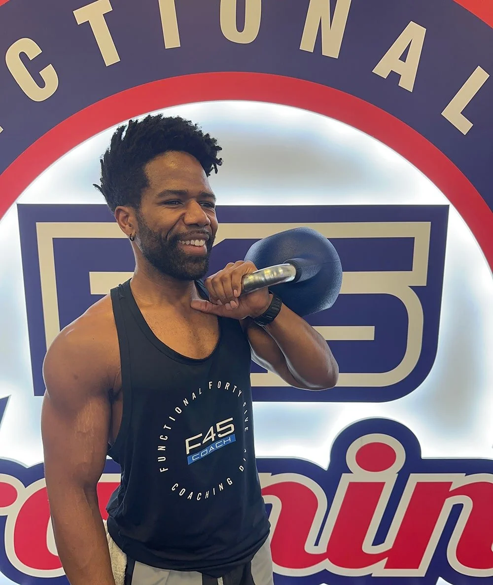 Man in a black athletic tank top lifting a kettlebell, smiling, in front of a sign that reads "National".