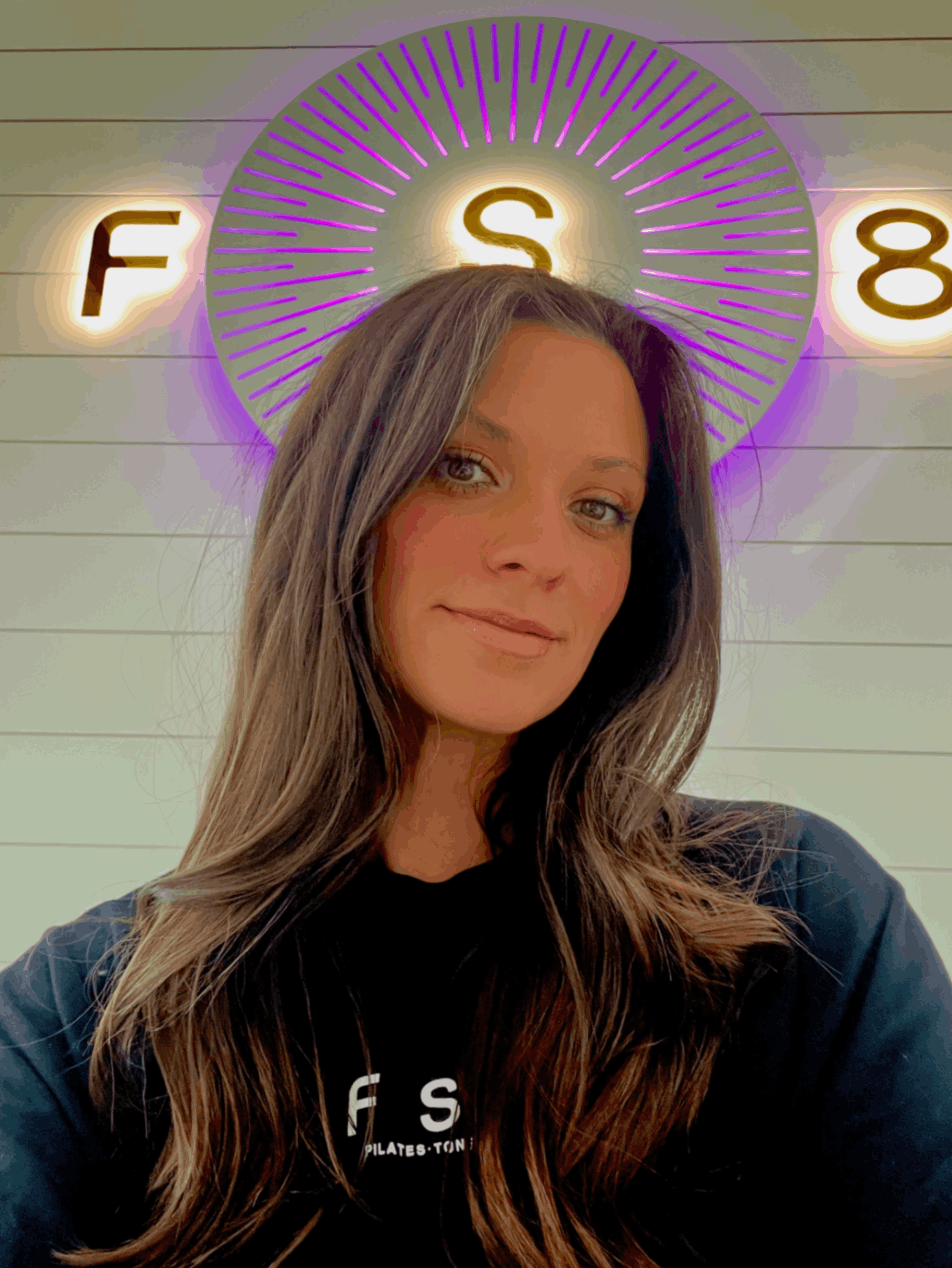 Young woman smiling and holding a yellow fitness hoop in front of a green wall.