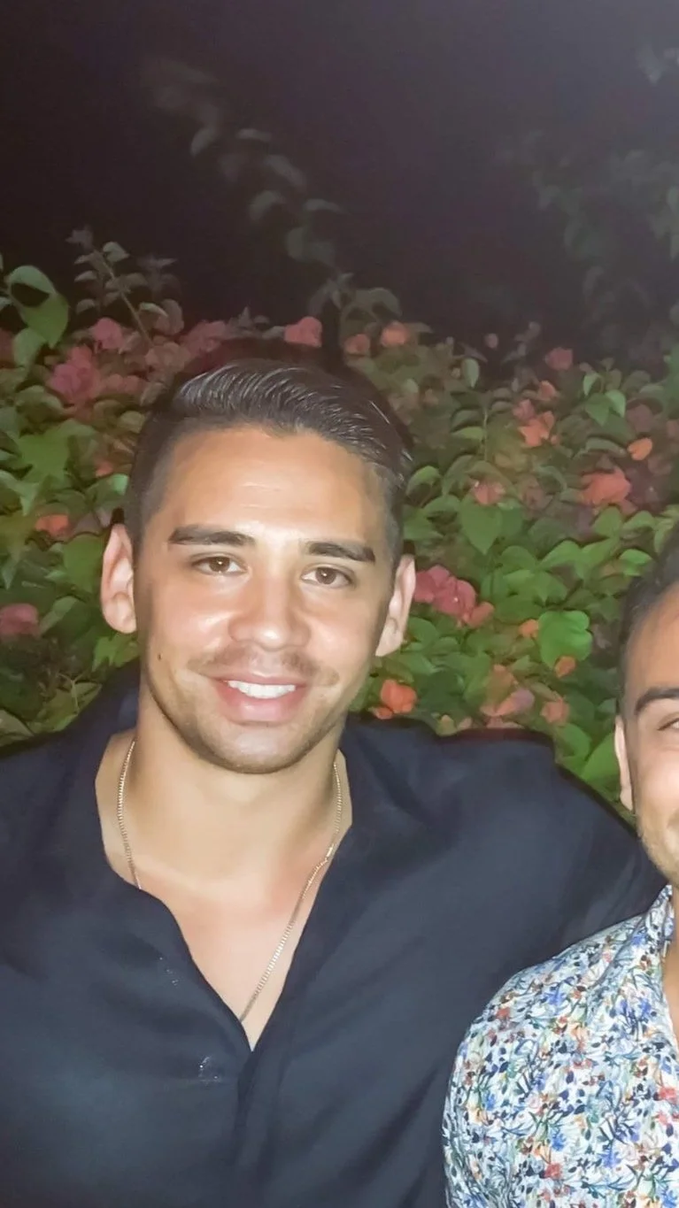 Young man with dark hair and beard wearing a white t-shirt standing against a light-colored wall.