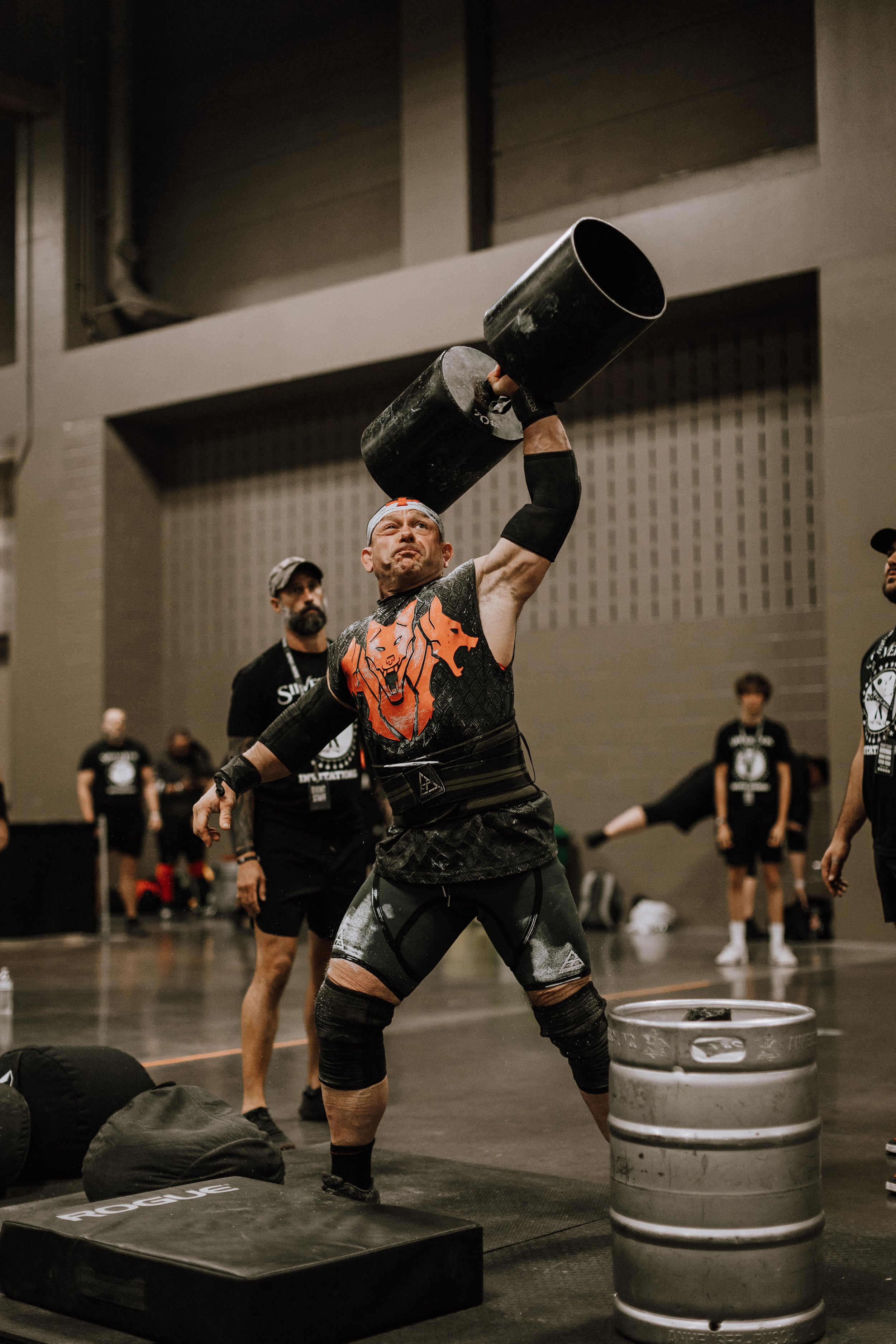 A man participating in a strongman competition is lifting a large, heavy black barrel over his head. He is wearing fitness gear, including knee sleeves and a shirt with a lion graphic. There are others in the background watching, and the setting appears to be an indoor gym or competition hall with various equipment and spectators.
