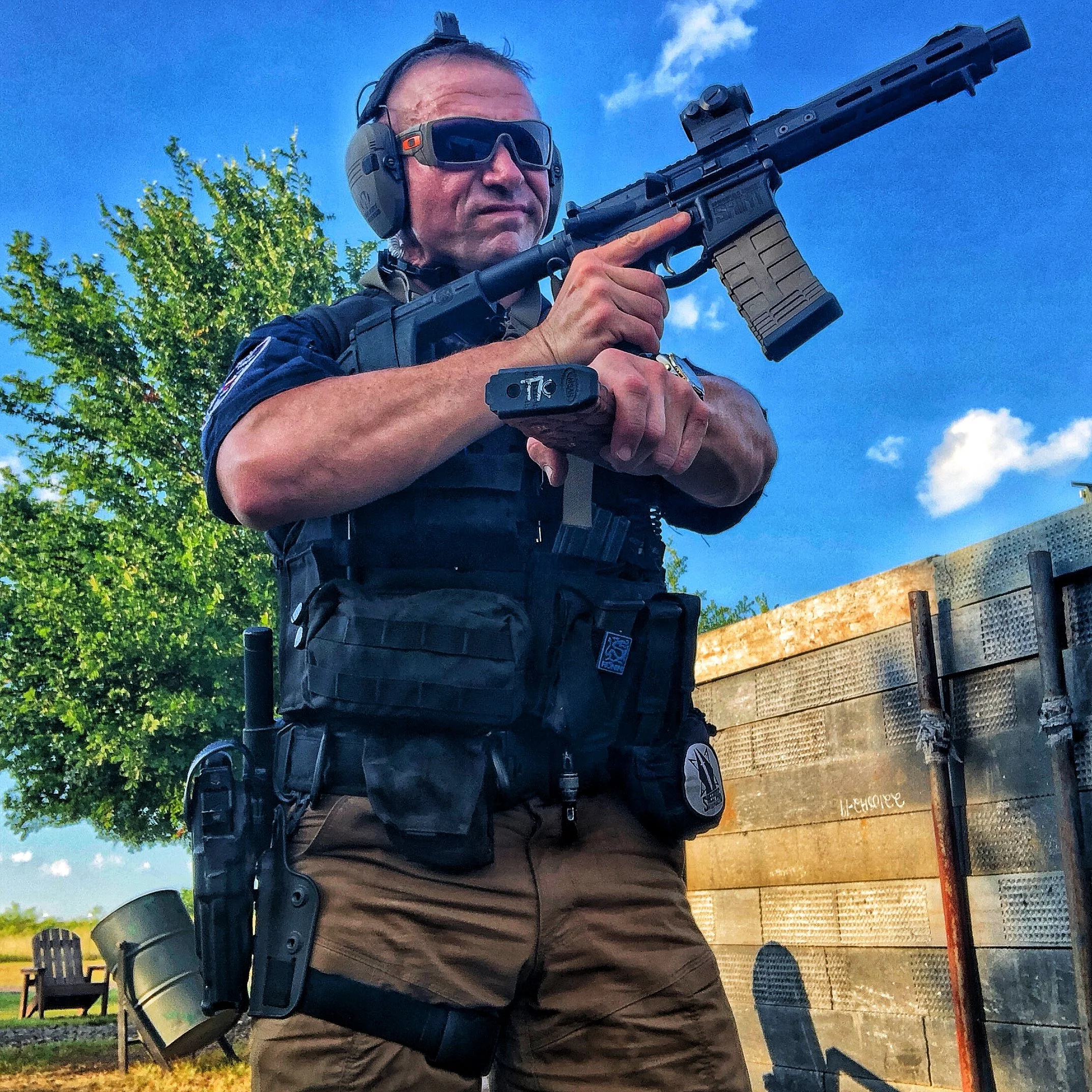 A man in tactical gear aiming a rifle outdoors against a blue sky and green trees.