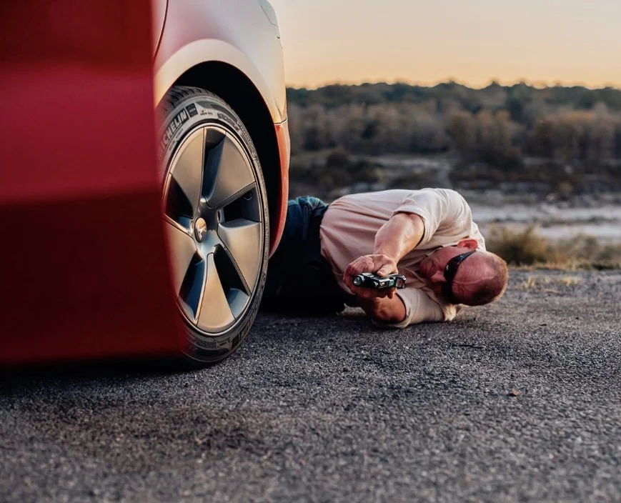 Man lying on the ground under a car, holding a remote control, in an outdoor setting at sunset.