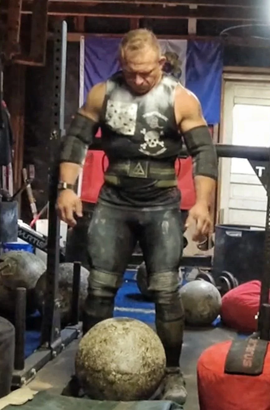 A muscular man in workout gear standing in a garage gym surrounded by weightlifting equipment and medicine balls.