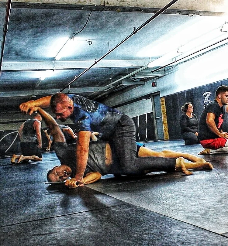 Two men practicing Brazilian jiu-jitsu on a black mat in a gym, one man on top pinning the other, with other people in the background sitting and kneeling.