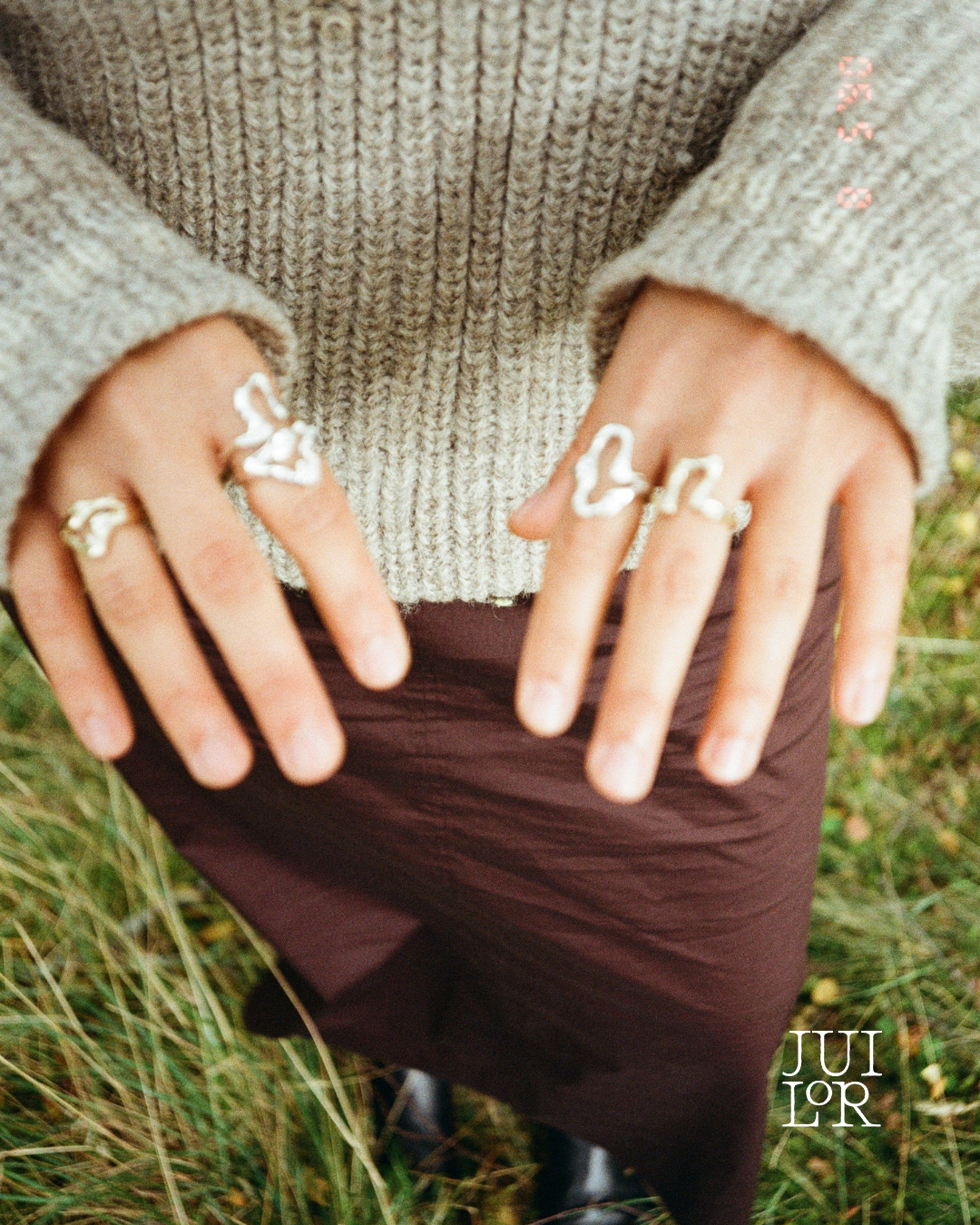 Close-up of hands wearing rings with white designs, wearing a beige knit sweater, with a grassy background.