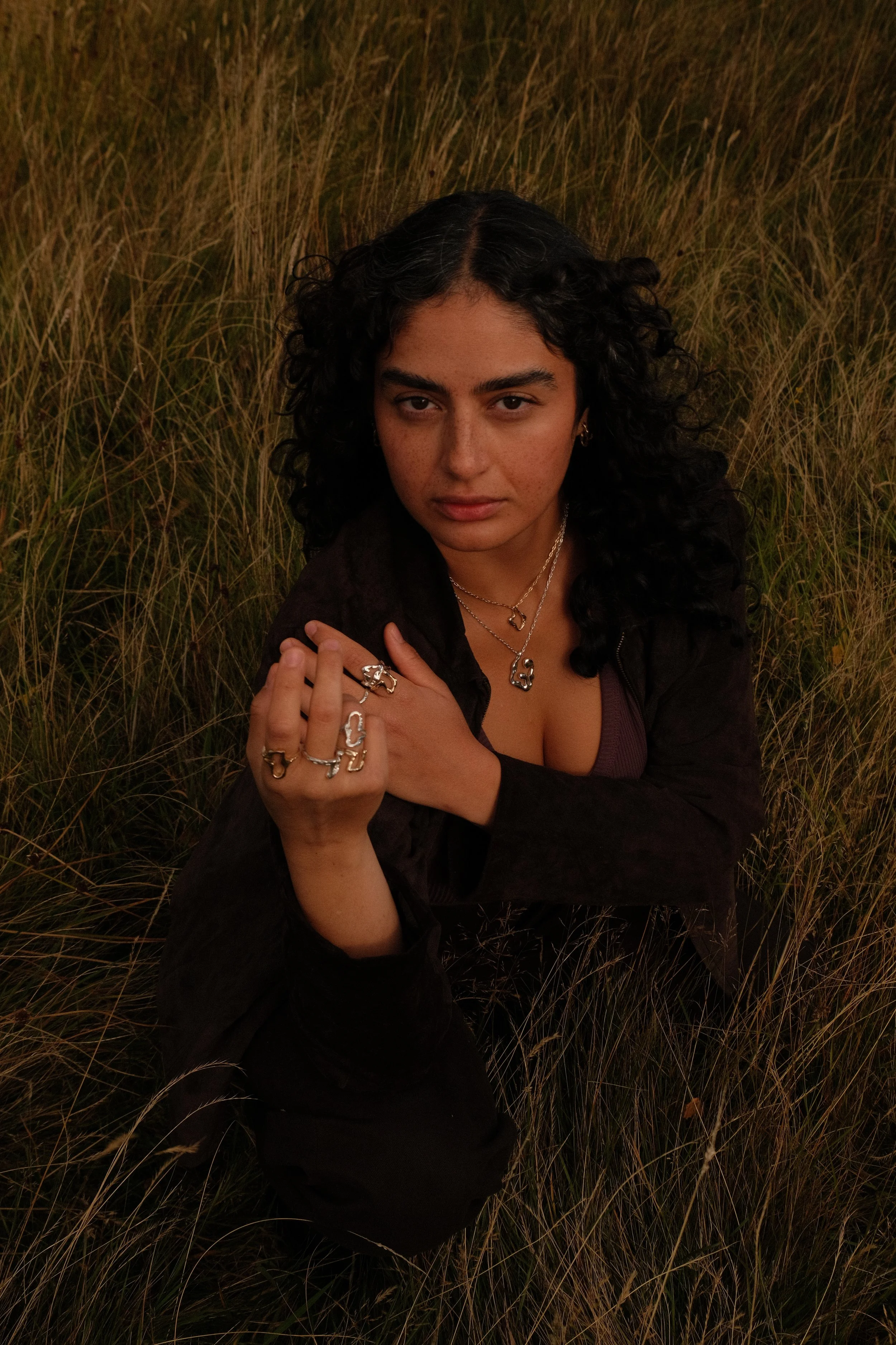 A woman with curly dark hair sitting in a grassy field during sunset, wearing multiple rings and necklaces, with a serious expression.