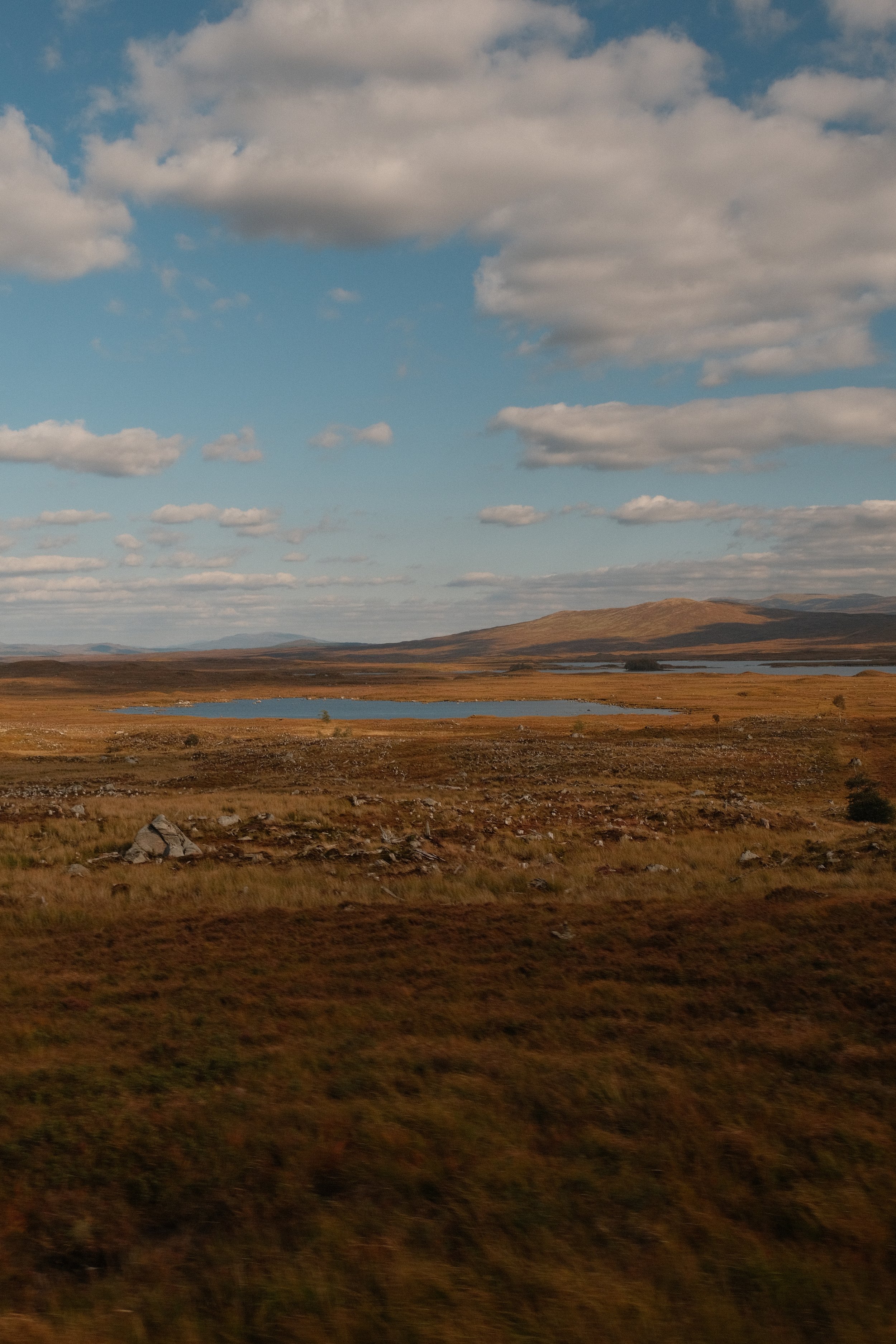 A wide landscape view of a grassy plain with a few small lakes, rolling hills, and mountains in the distance under a partly cloudy sky.