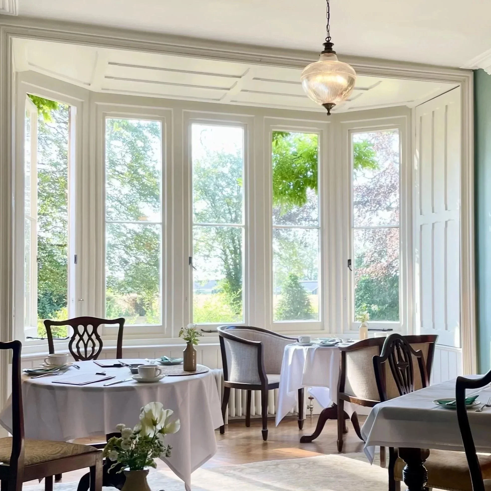 The Green's dining room with round tables set for a meal, large windows with greenery outside, and a vintage hanging light fixture.