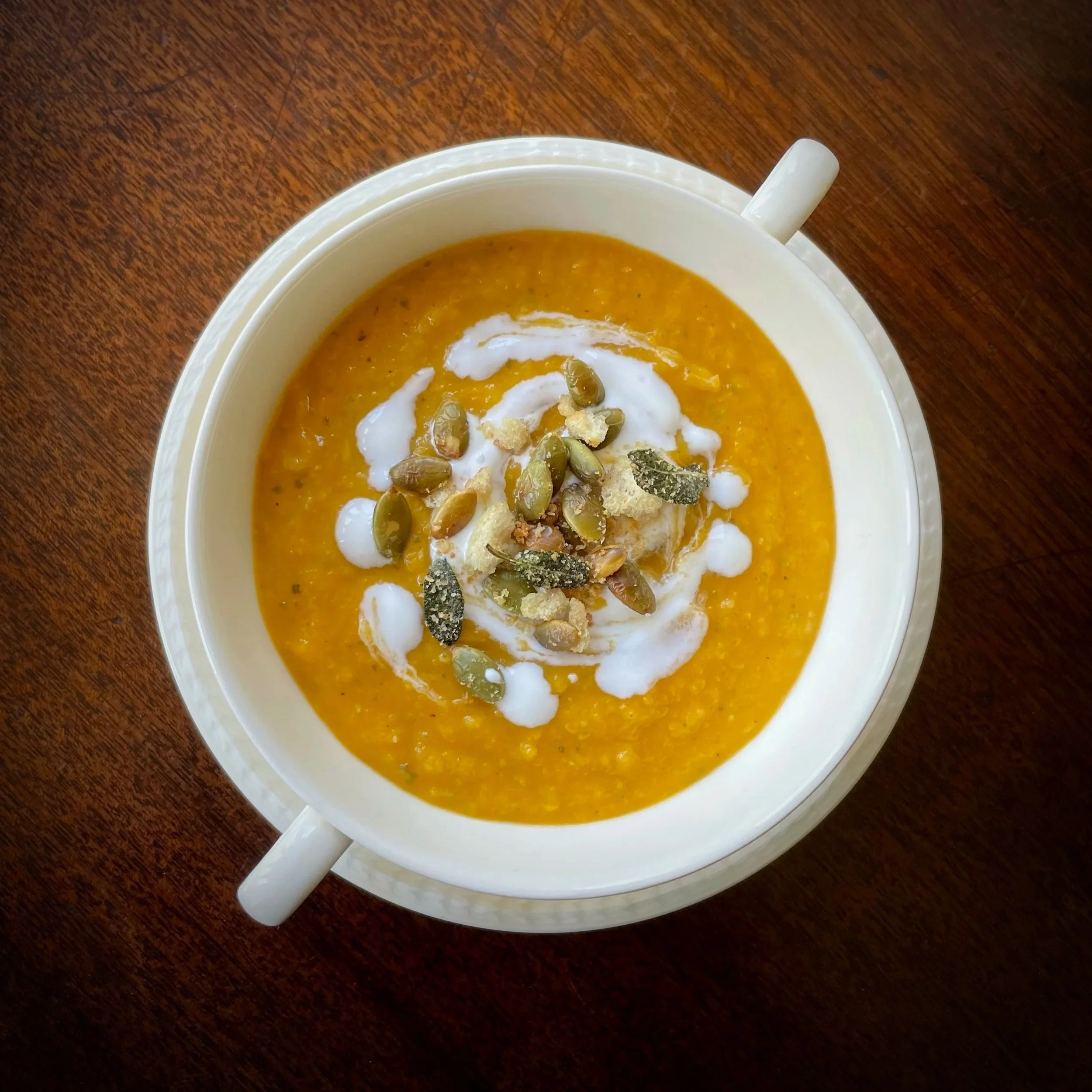 A bowl of orange-colored soup topped with white sour cream, pumpkin seeds, and herbs, placed on a wooden table.