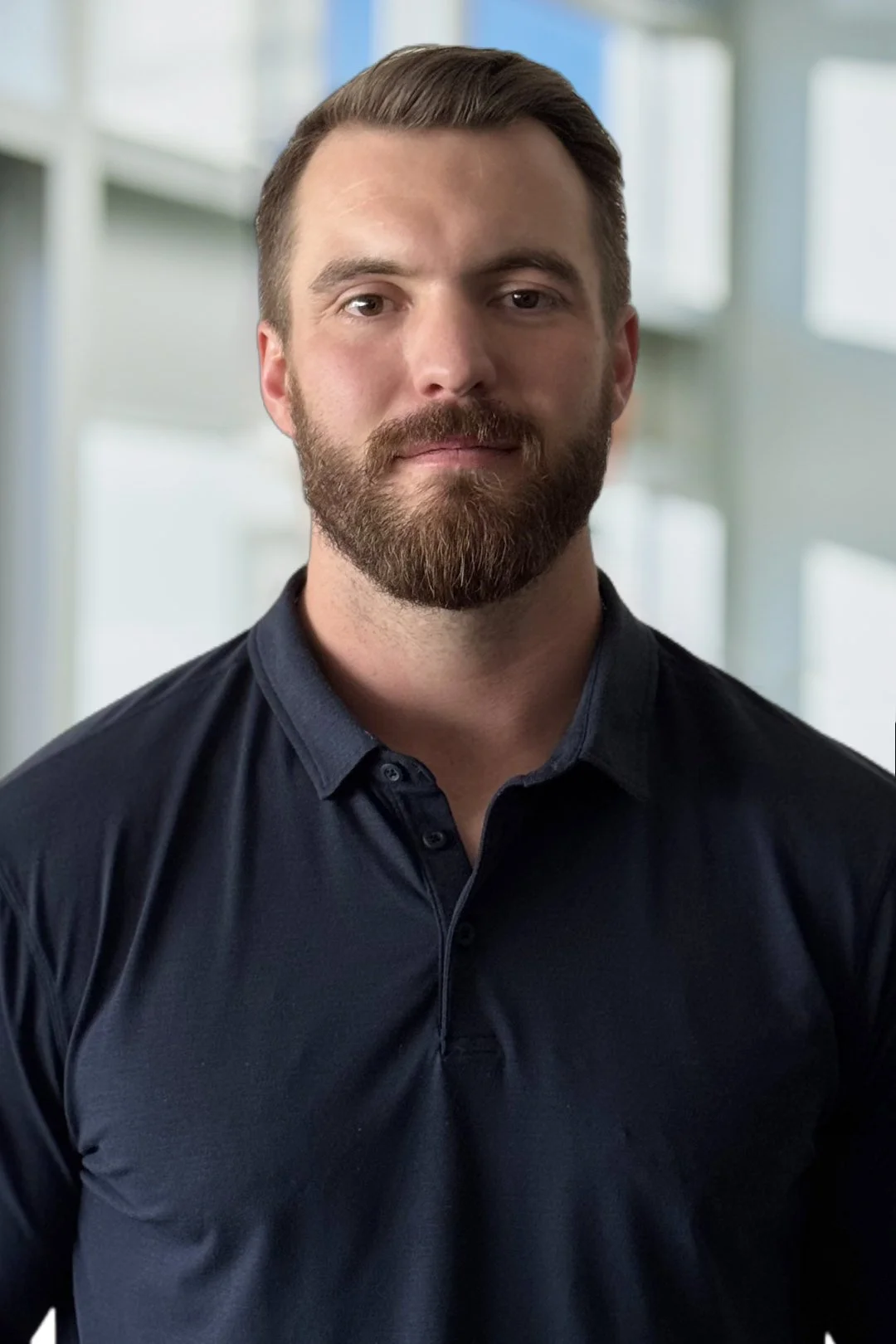 A man with a beard and short brown hair, wearing a dark navy button-up shirt, standing in front of a blurred modern office interior with large windows.
