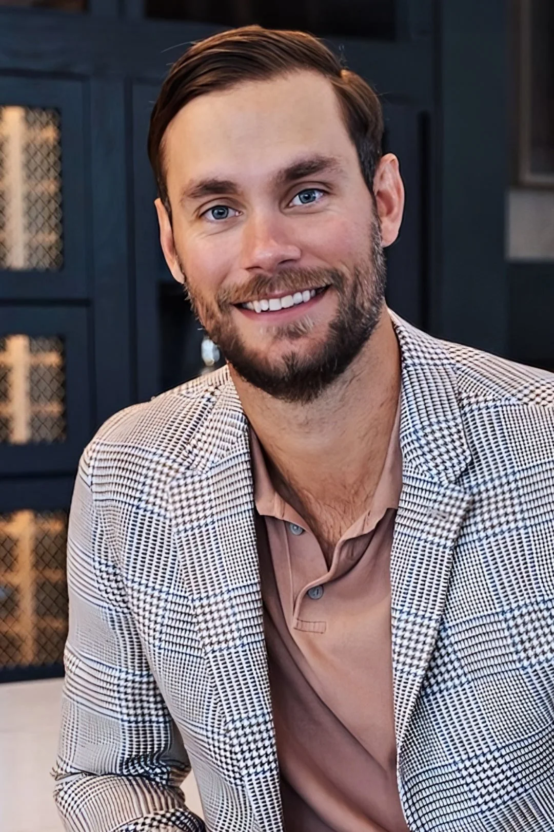 A smiling man with blue eyes, beard, and brown hair, wearing a checkered blazer and a beige dress shirt, sitting in a modern indoor setting.