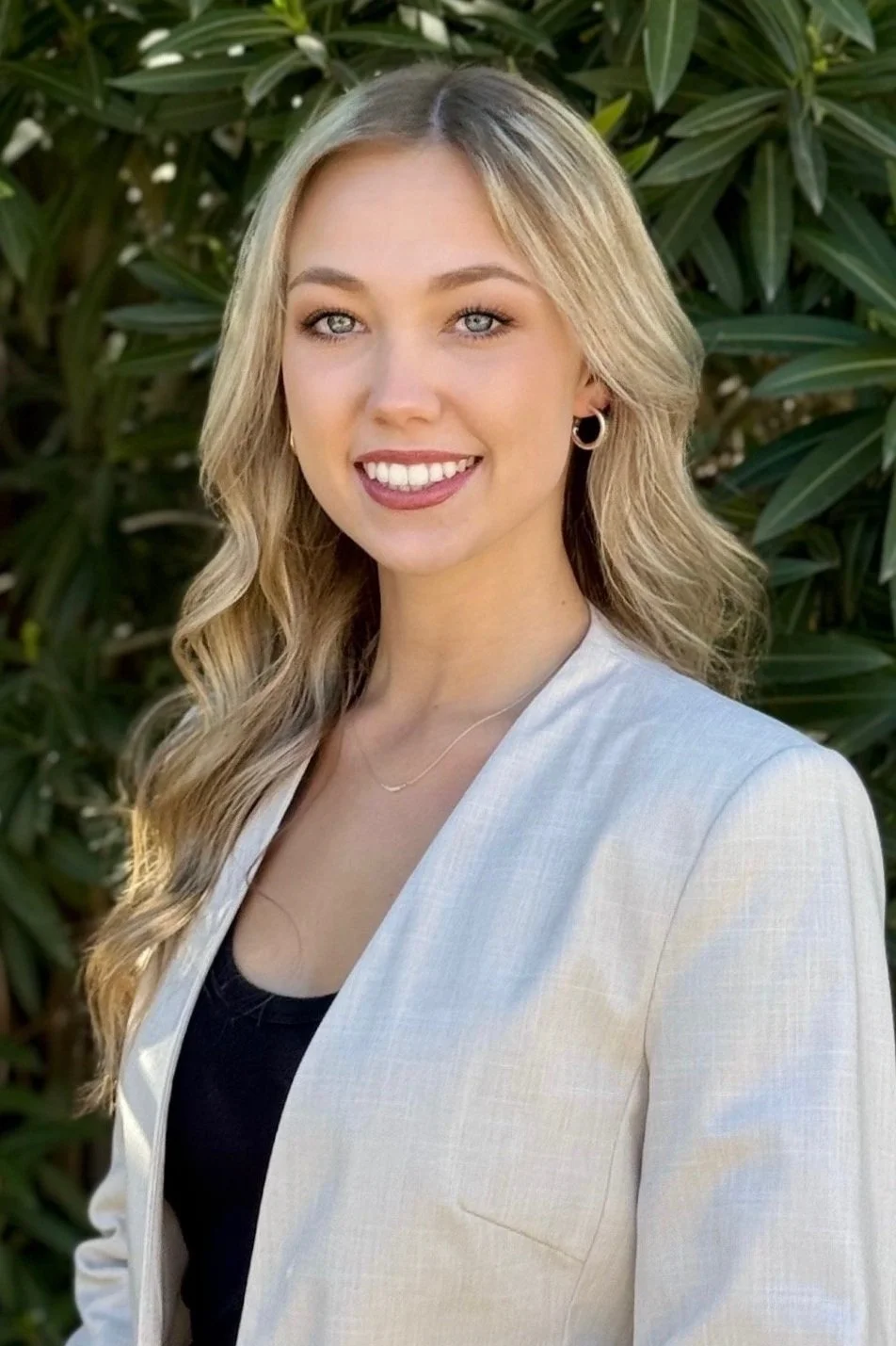 A young woman with blonde wavy hair, blue eyes, wearing earrings, a light-colored blazer, and a black top, standing outdoors with green foliage in the background.