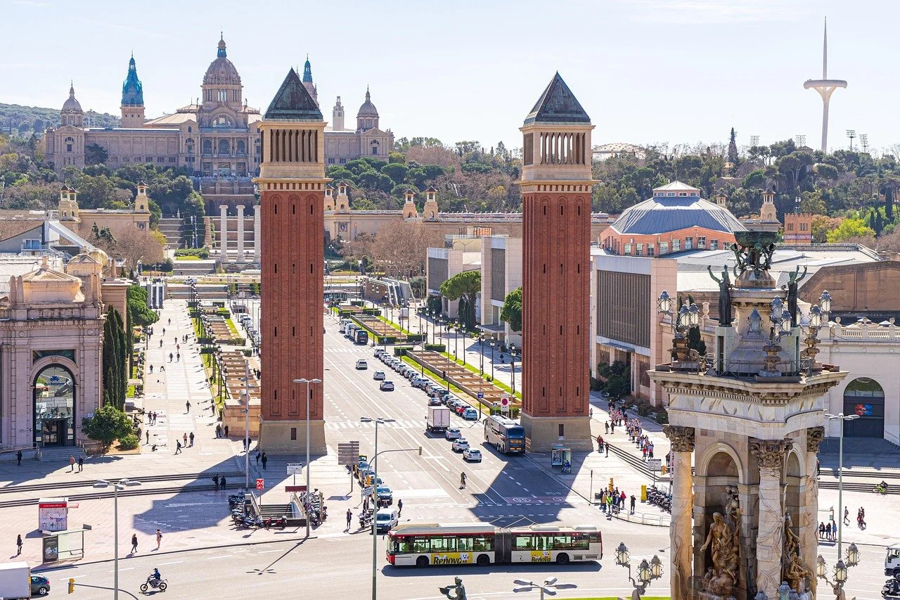 Street view with two red brick towers, busy intersection, people walking, buses, and historical buildings in the background, including a large domed building and a tower with a spire.
