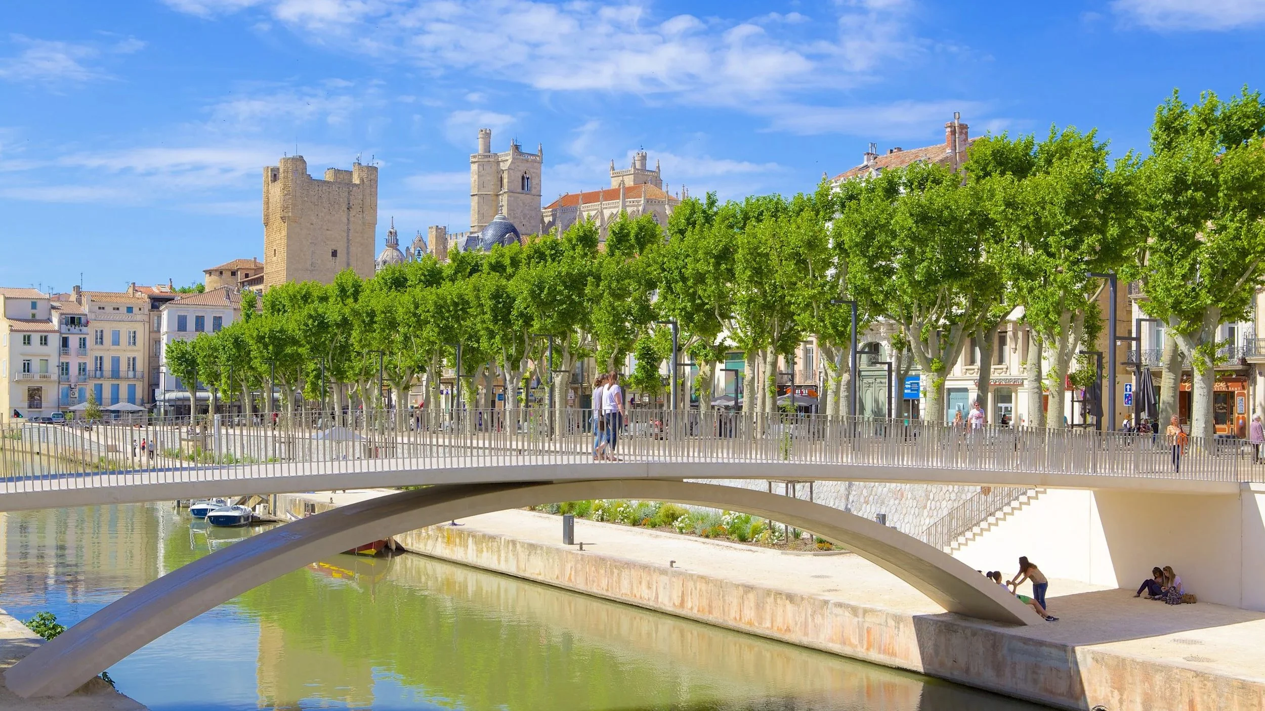 A cityscape featuring a river, a modern bridge with people walking, lush green trees lining the street, historic castle towers on a hill, and a bright blue sky with scattered clouds.