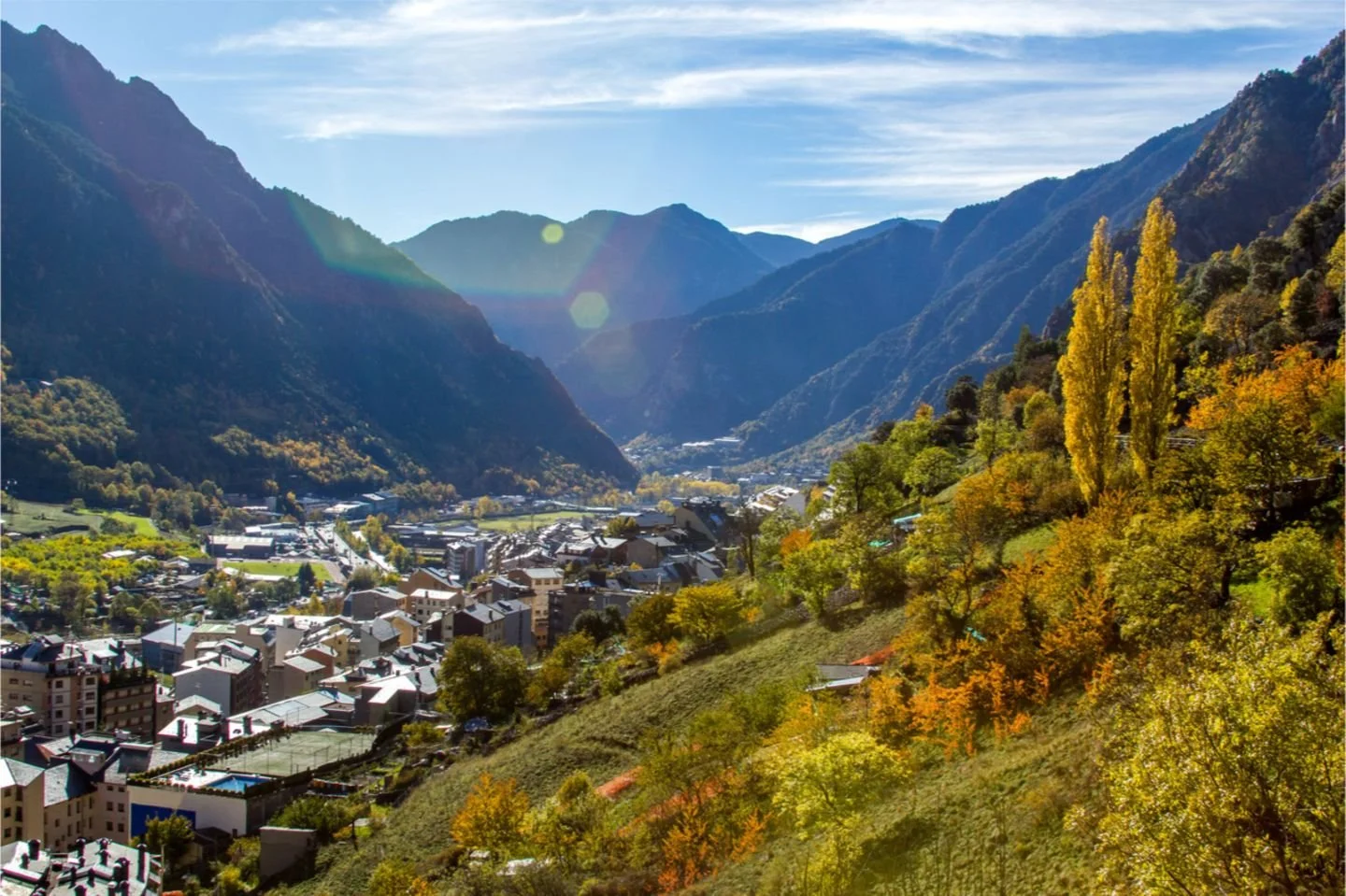 A mountainous valley with a small town and vibrant autumn-colored trees on the hillside, under a bright sky with visible lens flare.