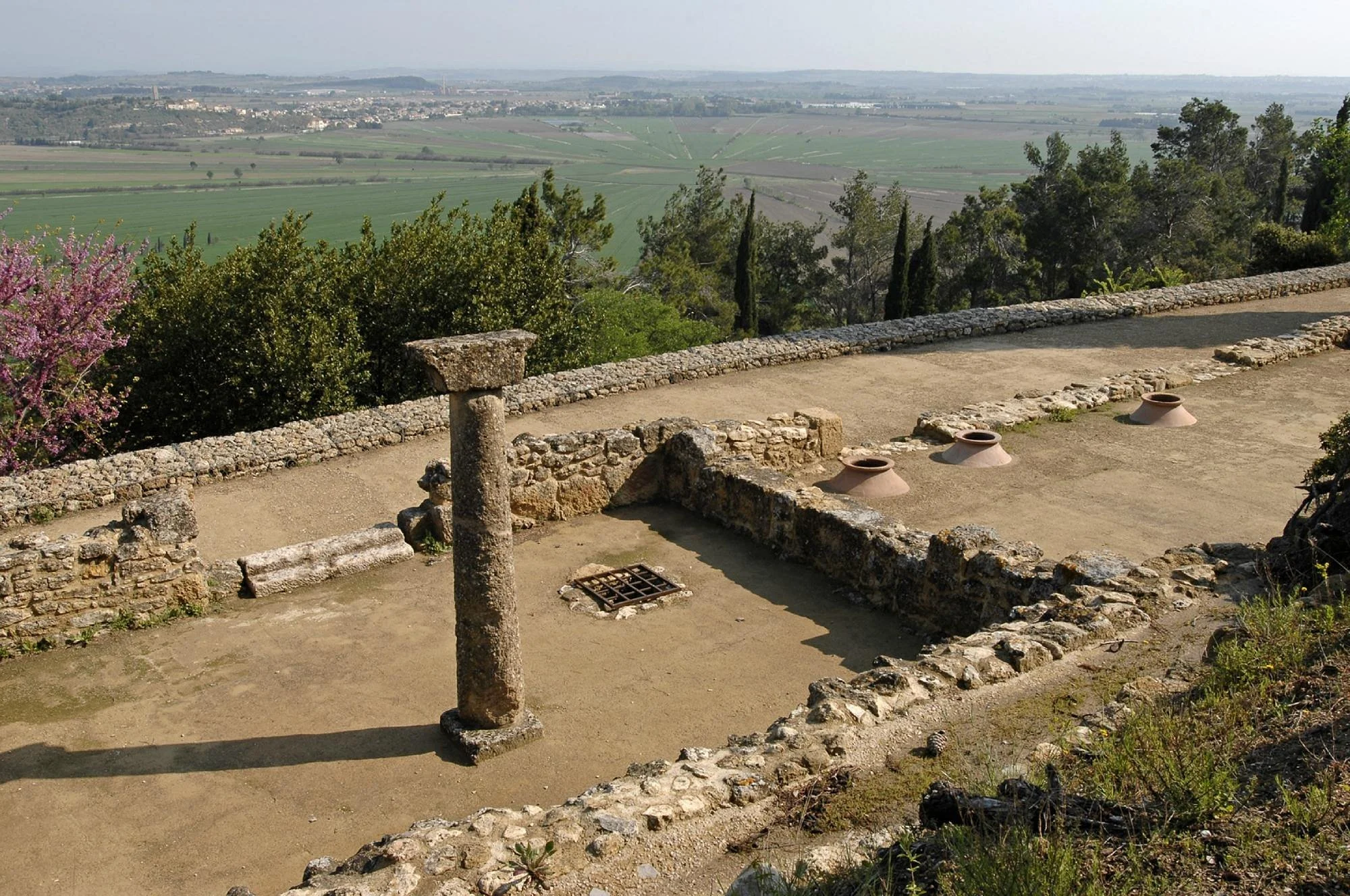 Archaeological site with stone structures, columns, and clay vessels overlooking a green landscape with trees and fields.