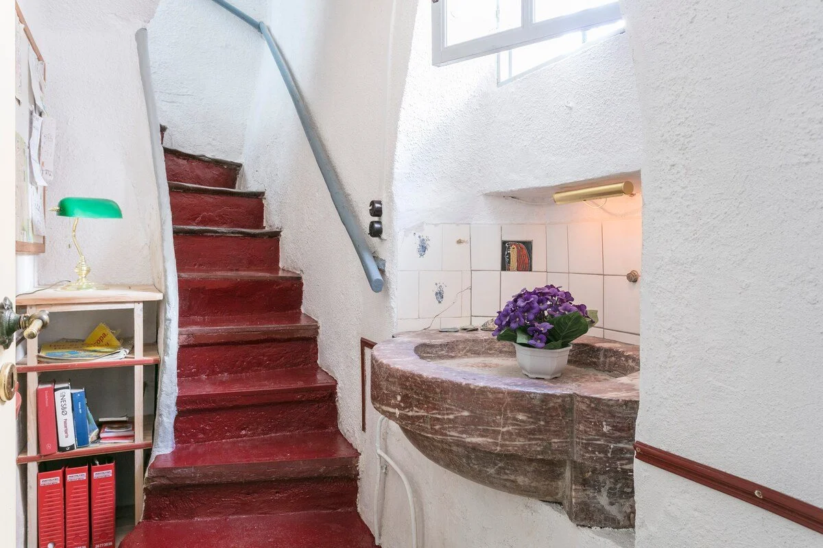 Indretning af et lille, rustikt rum med en rød trappe, der fører opad, en antik indbygget stensænk med lilla blomster, hvide strukturerede vægge og et vindue, der lukker naturligt lys ind.