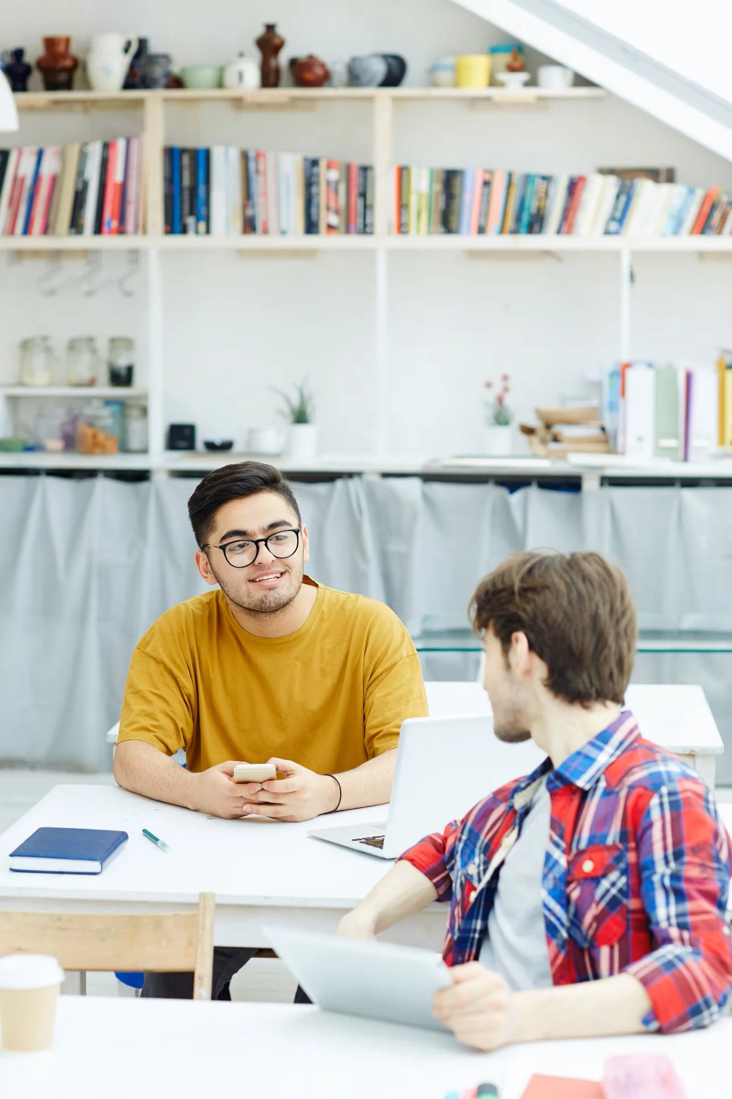 Two young men having a conversation at a white table in a bright, modern room. One is wearing a yellow shirt and glasses, the other a checkered shirt. They have laptops, notebooks, and a coffee cup on the table.