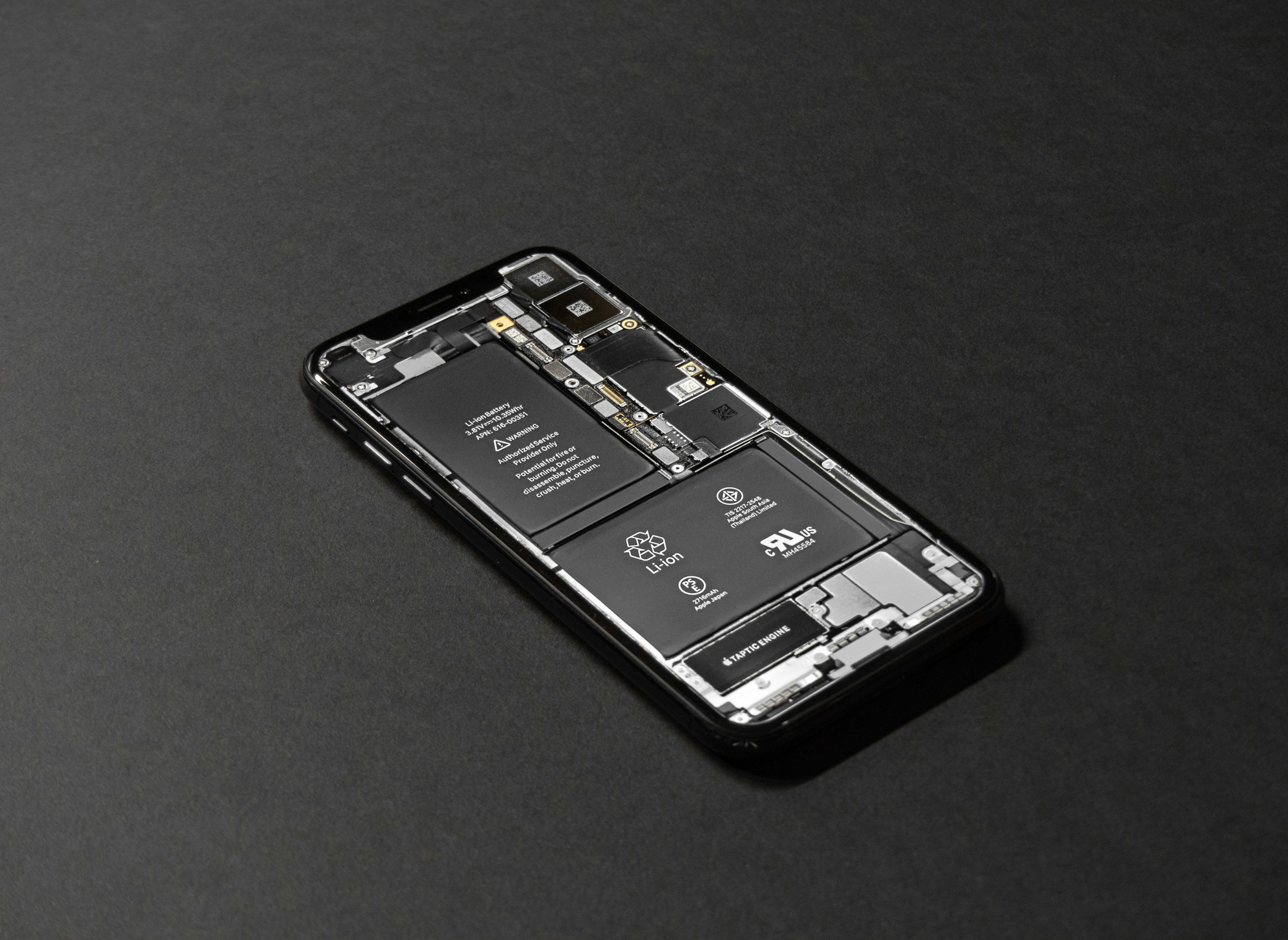 A modern smartphone with its back cover removed, revealing internal components such as batteries, circuit boards, and chips, placed on a dark surface.