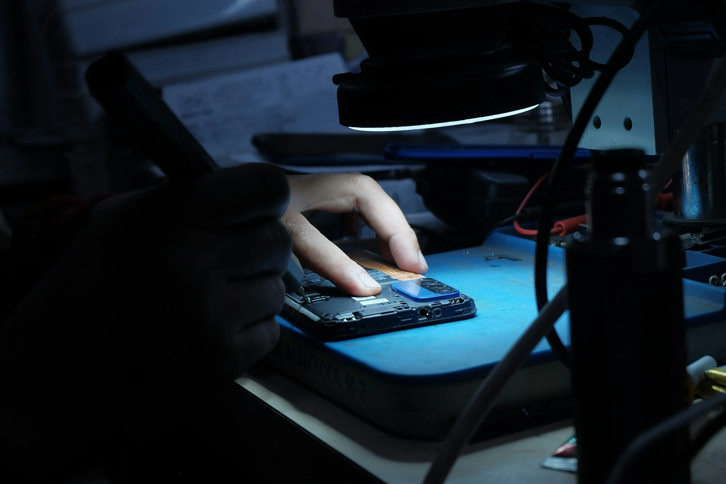 Close-up of a person repairing a smartphone under a lamp on a workbench.