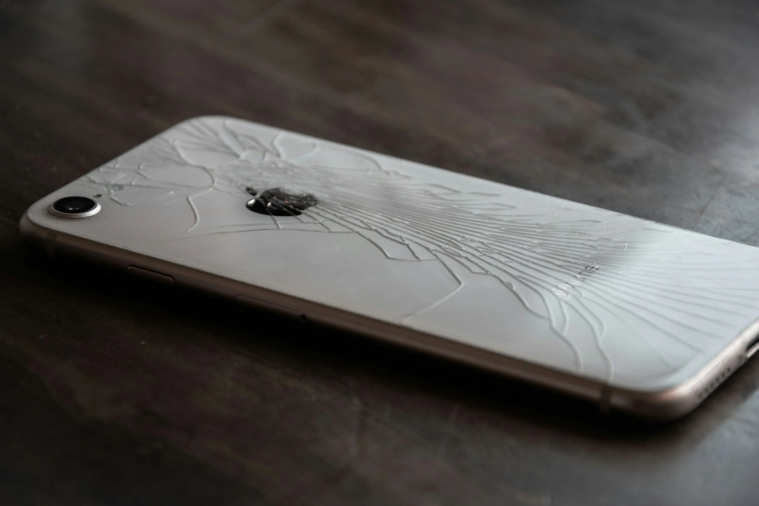 A white smartphone with a shattered back glass lying on a dark wooden surface.