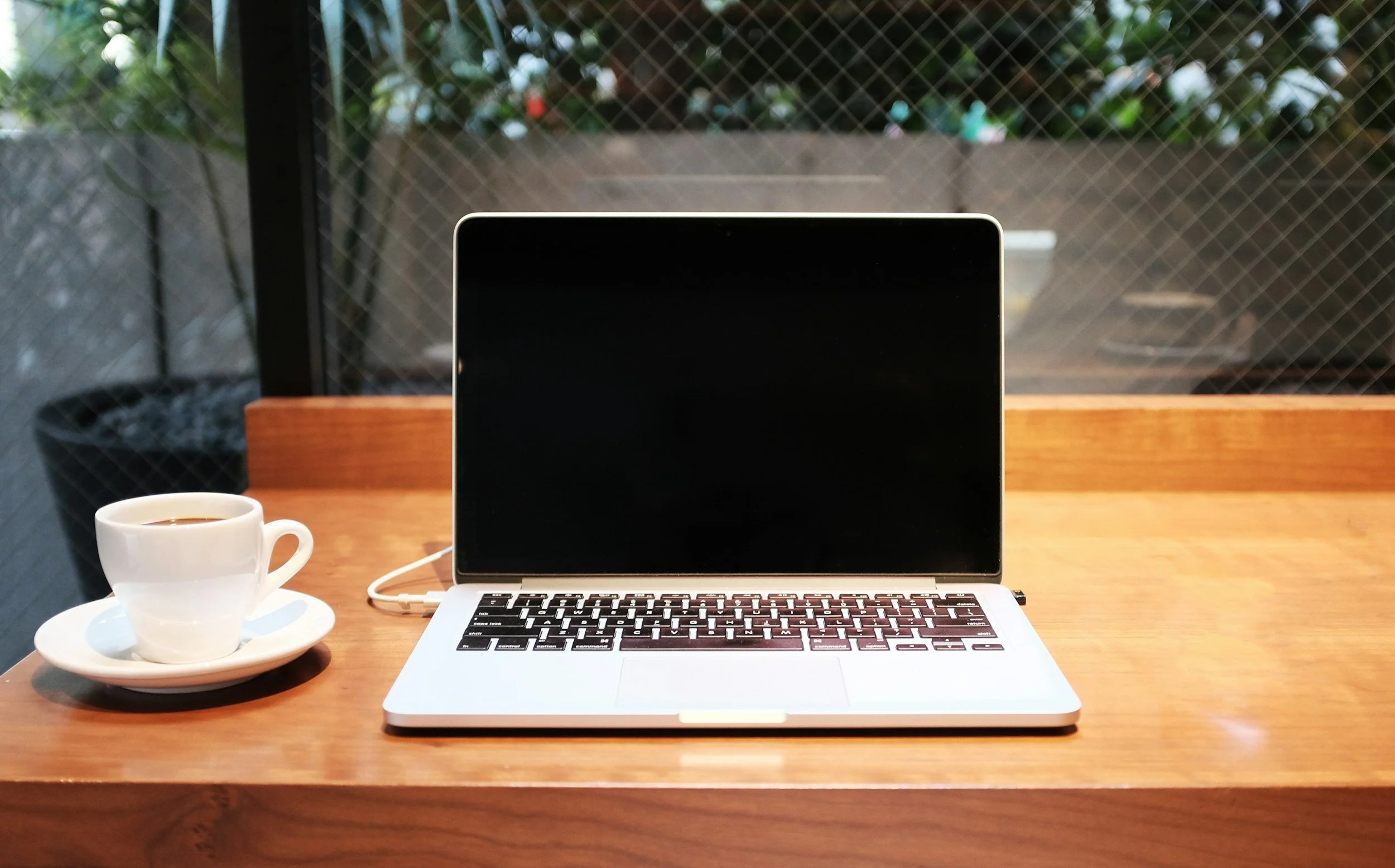 Open laptop computer on a wooden table next to a white coffee cup with black coffee, against a window with a mesh screen and outdoor plants in the background.