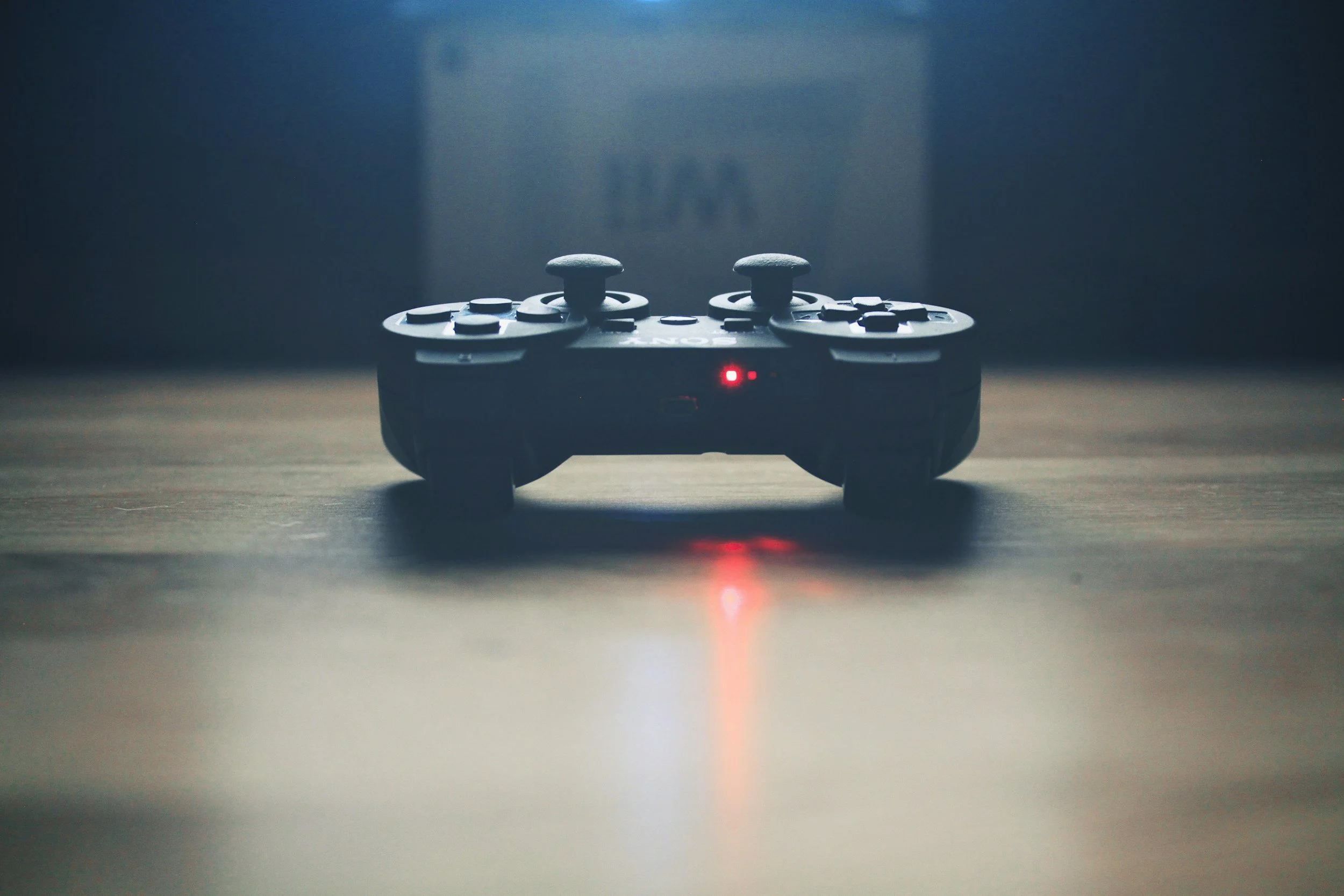 A black gaming controller with a red LED light, placed on a wooden surface, in a dimly lit room.