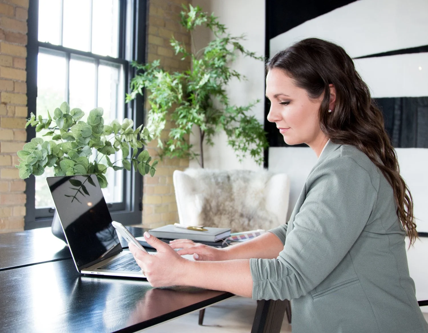Alicia sitting at a desk using a smartphone with a laptop and books in front of her, in a room with large windows, green plants, and brick and white walls.