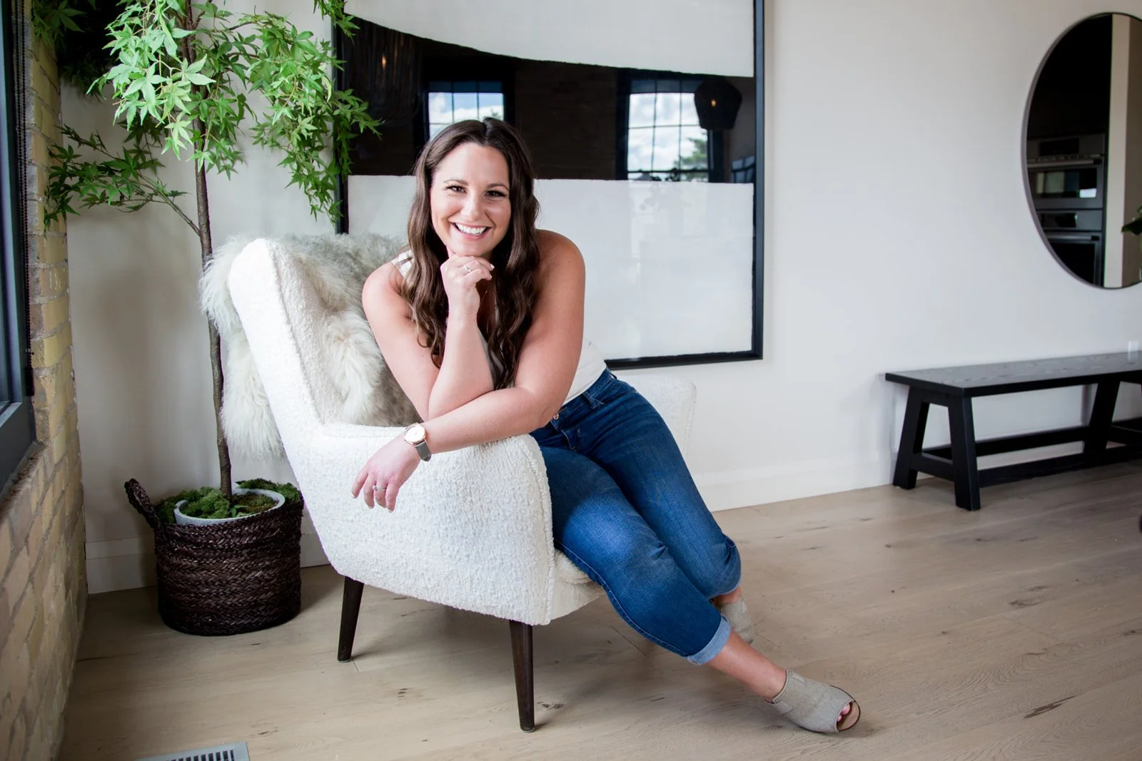 Alicia, founder of Boulevard North, Web and Creative, sitting on a white textured armchair, smiling, in a modern room with a large window showing a blue sky, a round mirror on the wall, and a potted plant nearby.