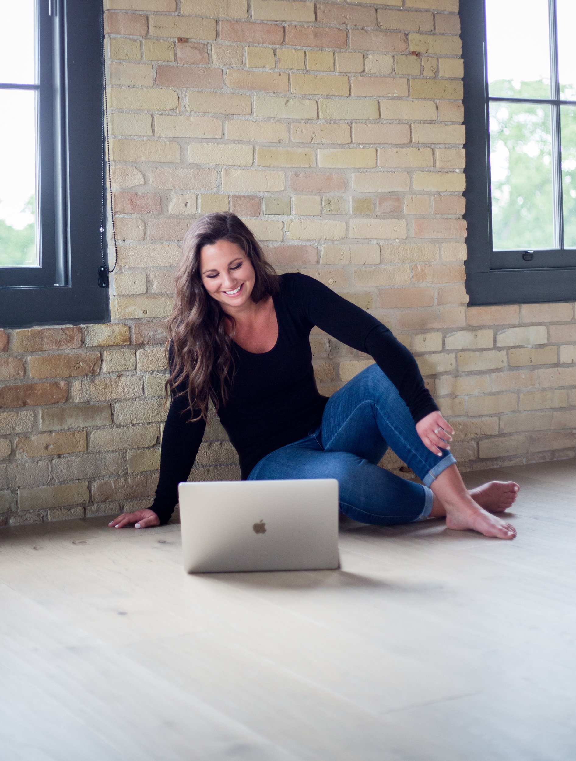 Alicia, founder of Boulevard North, Web and Creative, sitting on a wooden floor with a silver MacBook laptop in front of her, smiling, in a room with brick walls and large windows. Planning out her logo project with a happy client.