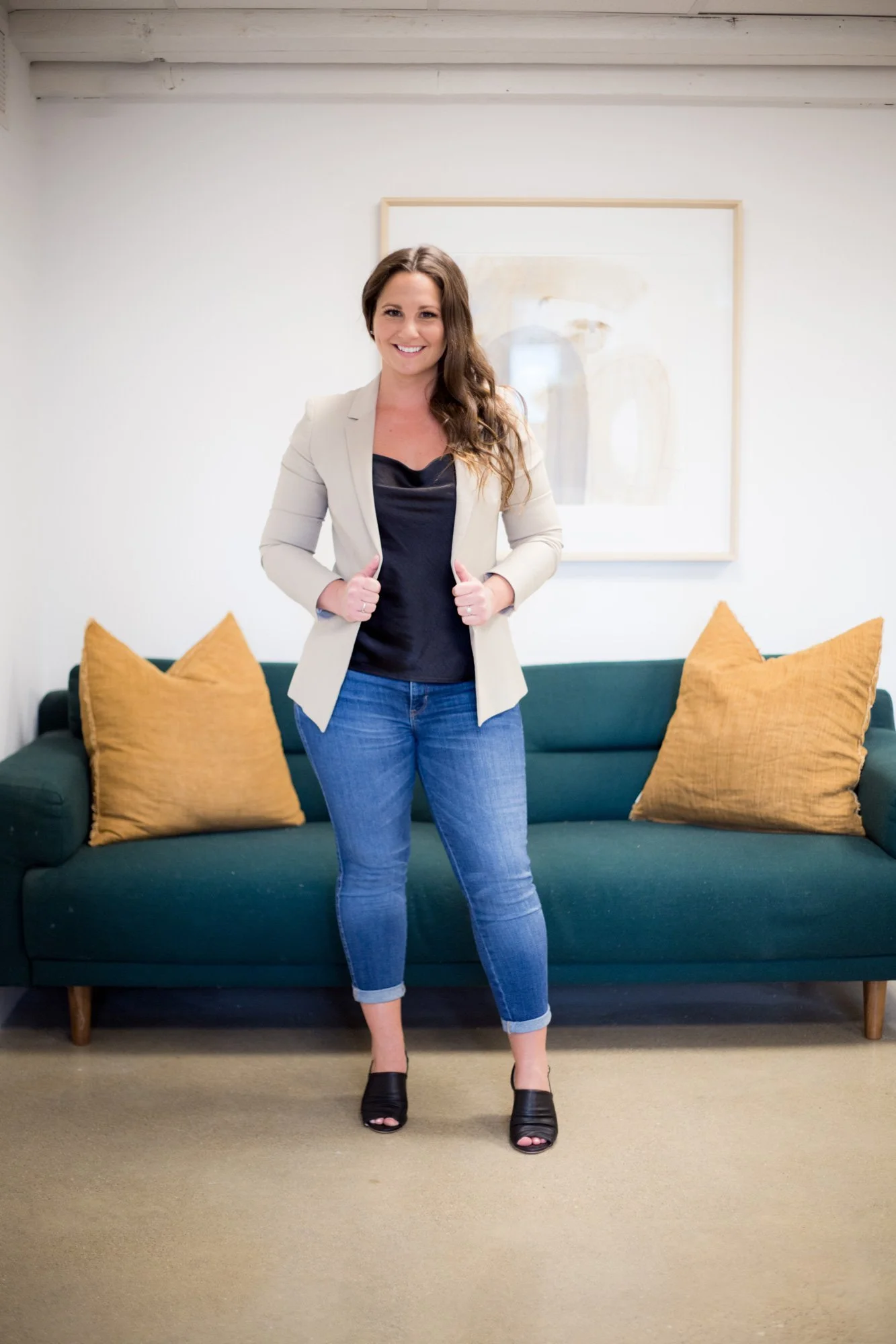 Alicia standing in front of a green sofa with two mustard yellow pillows, wearing a beige blazer, black top, blue jeans, and black heeled shoes, in a room with a minimalistic white wall and artwork.