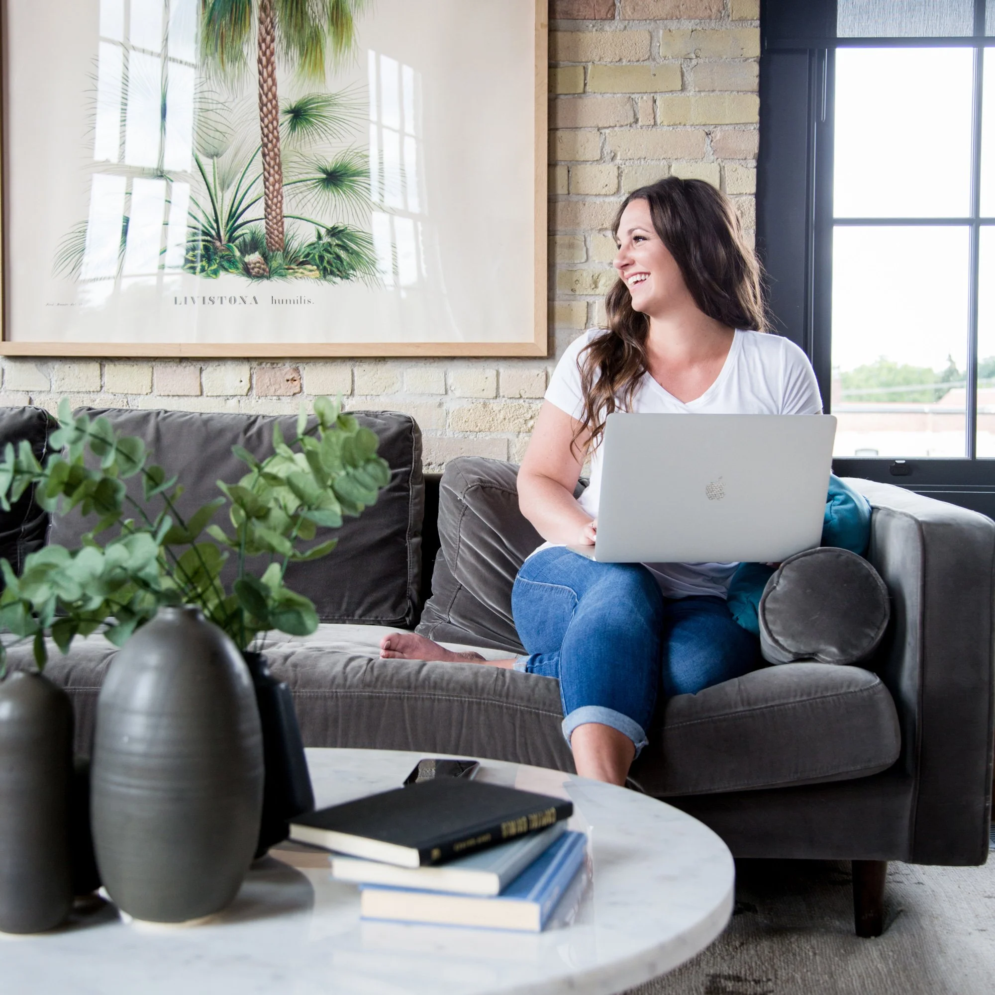 Alicia, owner of Boulevard North, sitting on a grey couch with a laptop on her lap, smiling and looking to her right in a modern living room with a brick wall, large window, and framed artwork of a plant. Designing a website and logo for a client.