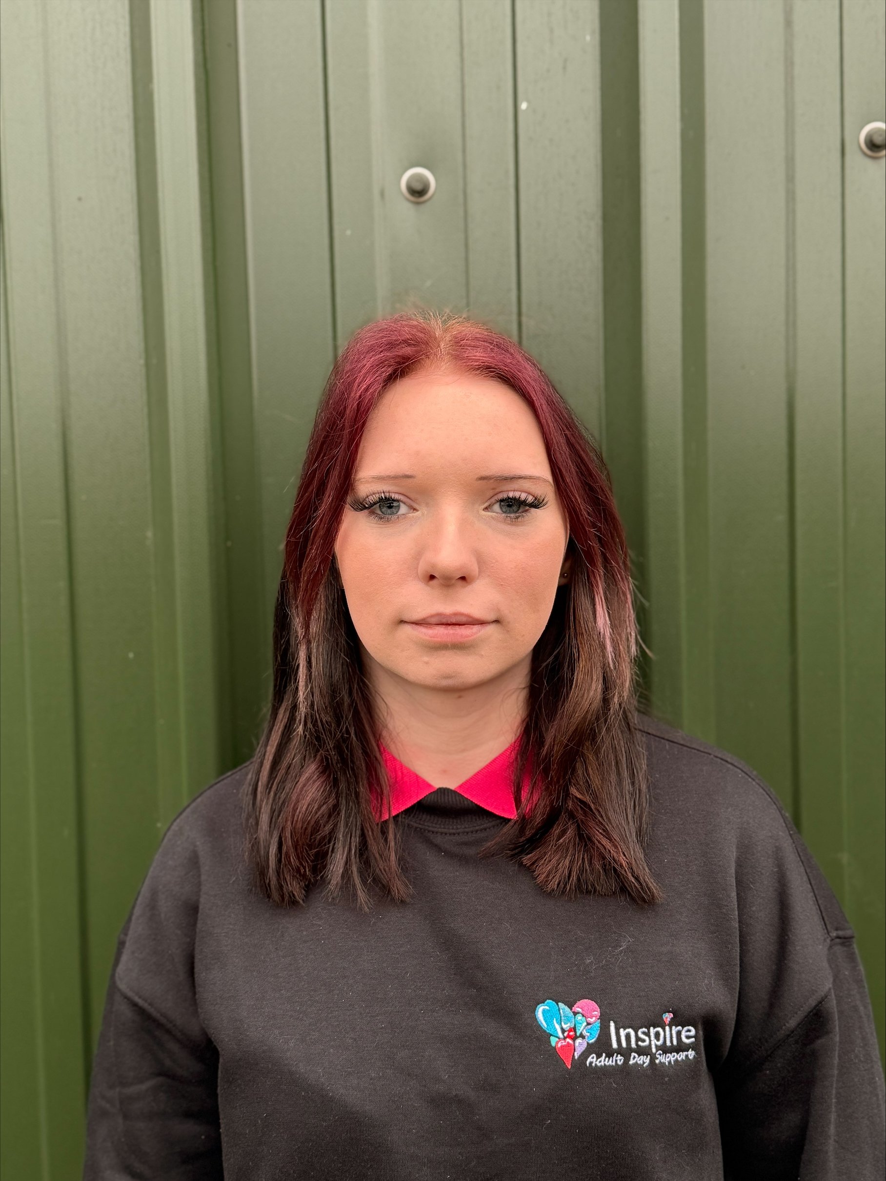 A smiling middle-aged woman with short hair, wearing a pink polo shirt with the logo 'Inspire Adult Day Support,' standing outdoors with green foliage in the background.