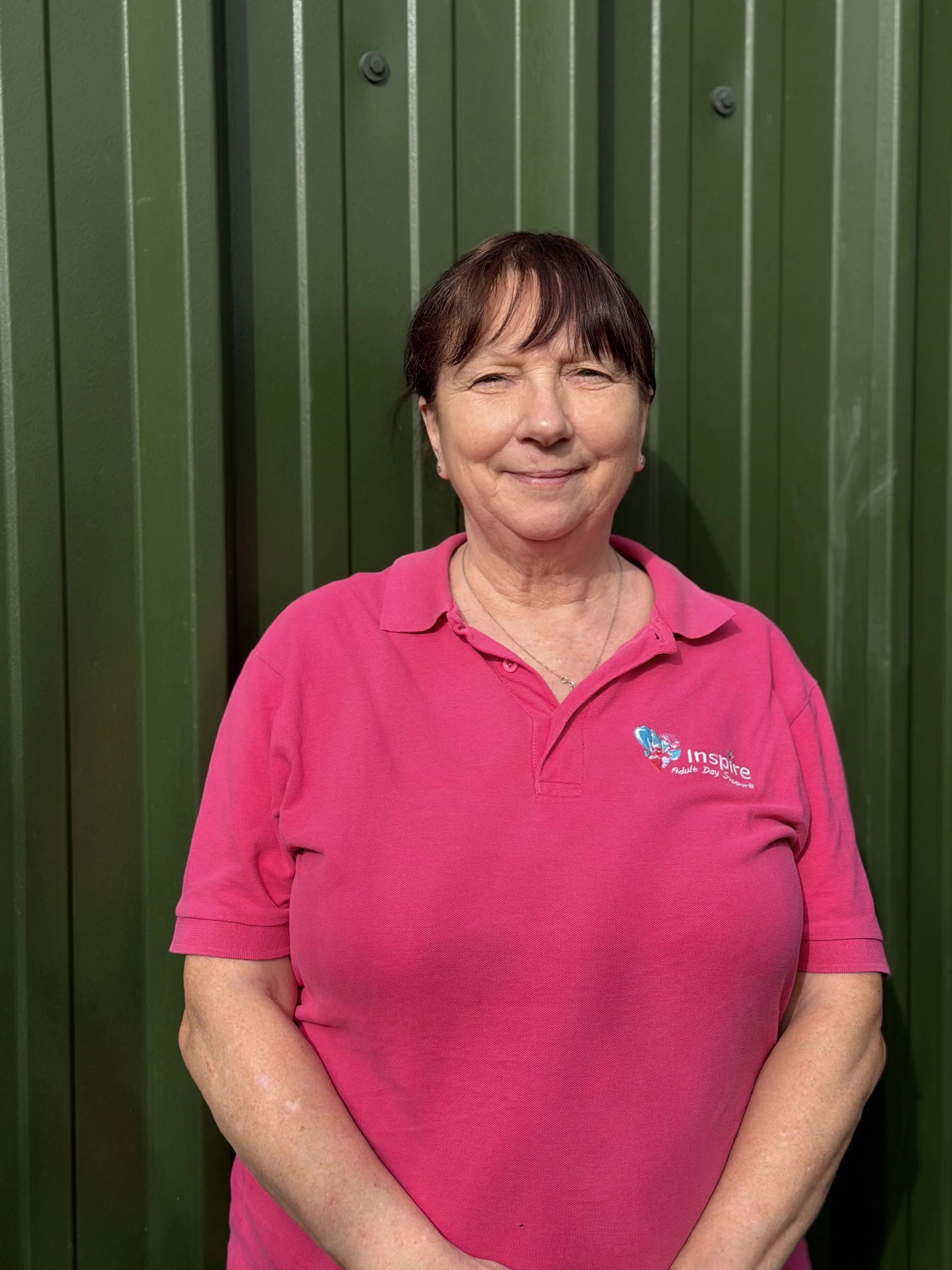 A smiling middle-aged woman with short hair, wearing a pink polo shirt with the logo 'Inspire Adult Day Support,' standing outdoors with green foliage in the background.