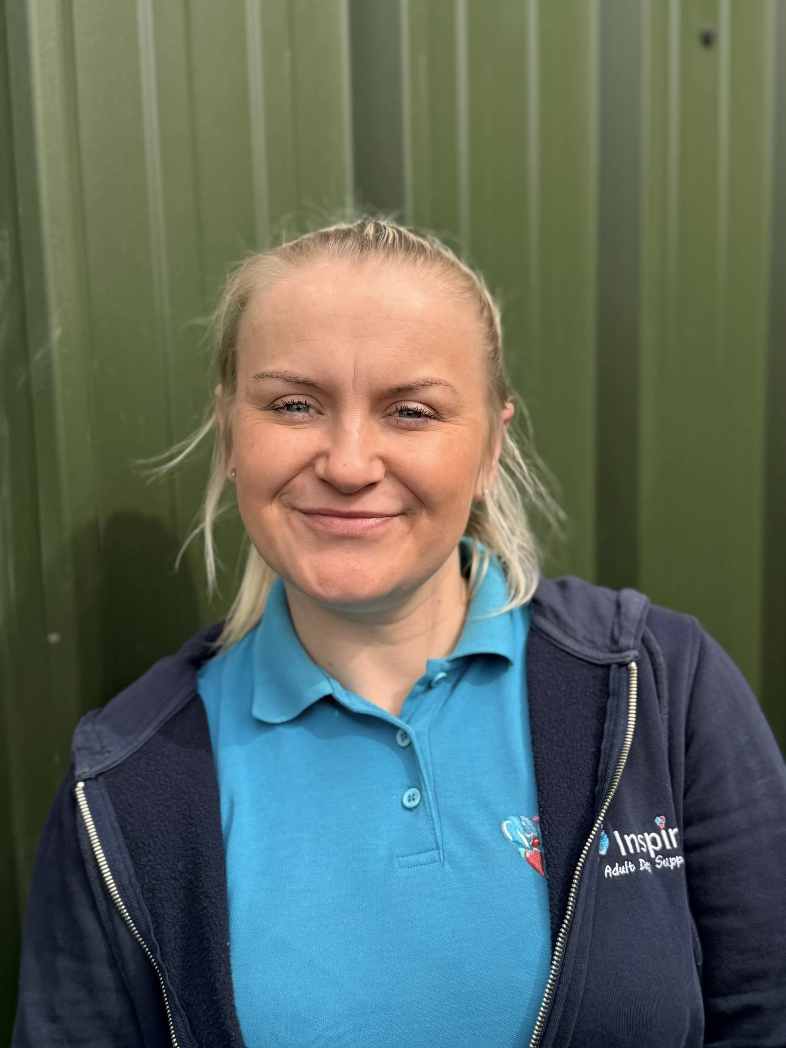 A woman outdoors wearing a blue polo shirt with an 'Inspire Adult Day Support' logo, smiling with closed eyes, background of green foliage.