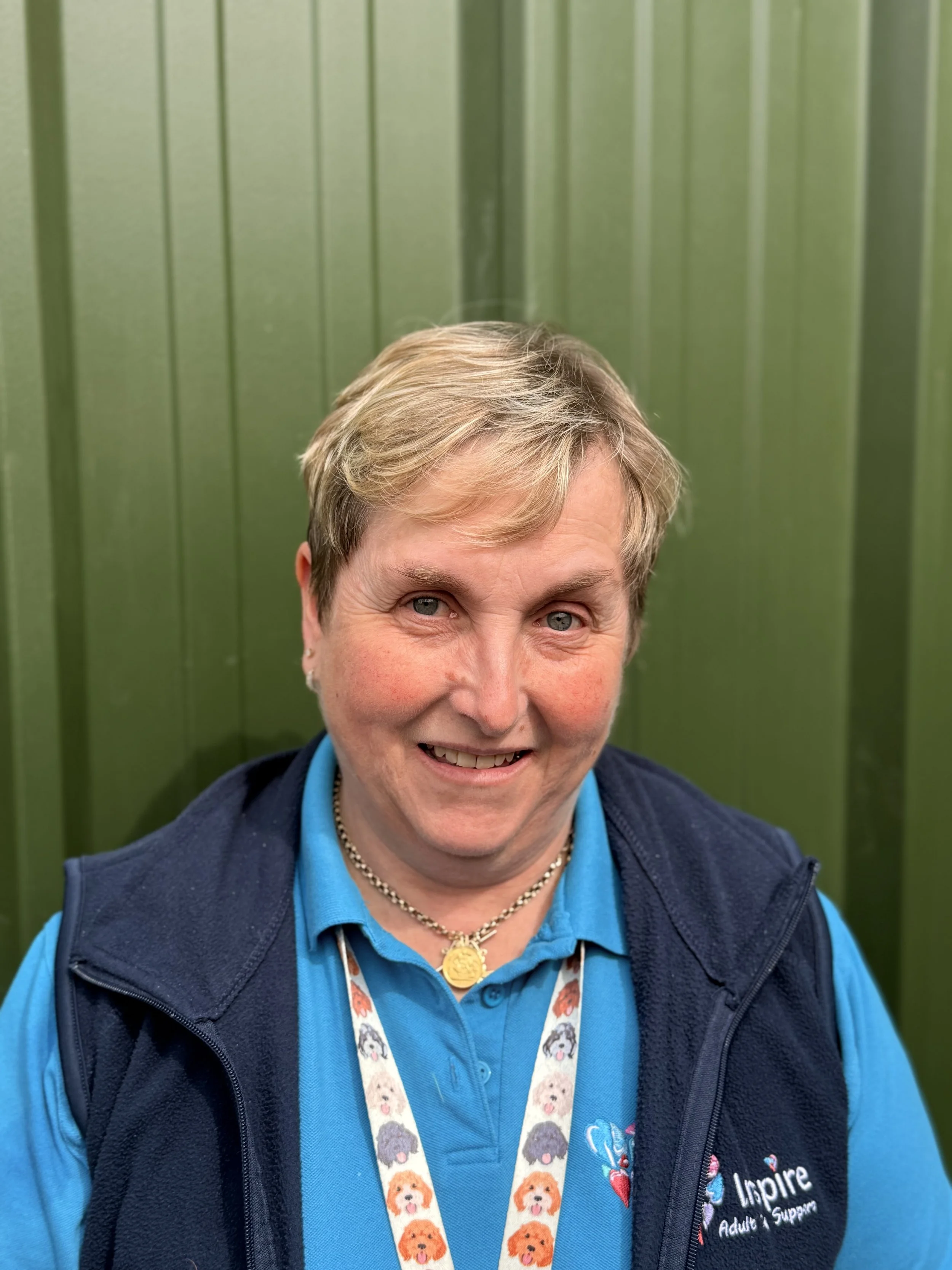 A smiling middle-aged woman with short hair, wearing a pink polo shirt with the logo 'Inspire Adult Day Support,' standing outdoors with green foliage in the background.