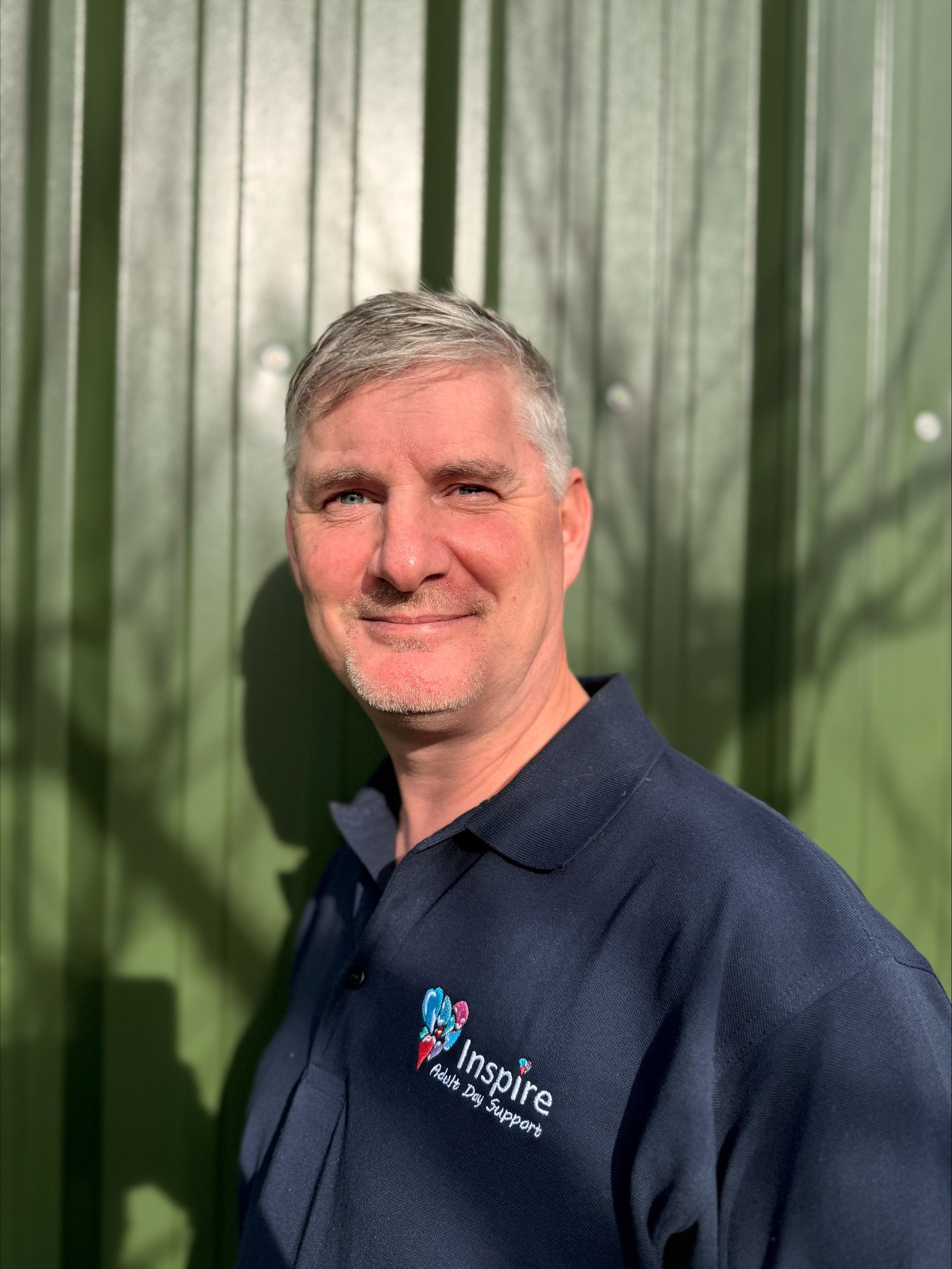 A woman with blonde hair pulled back, wearing a teal polo shirt and a blue staff lanyard, standing outdoors in front of leafy green bushes.
