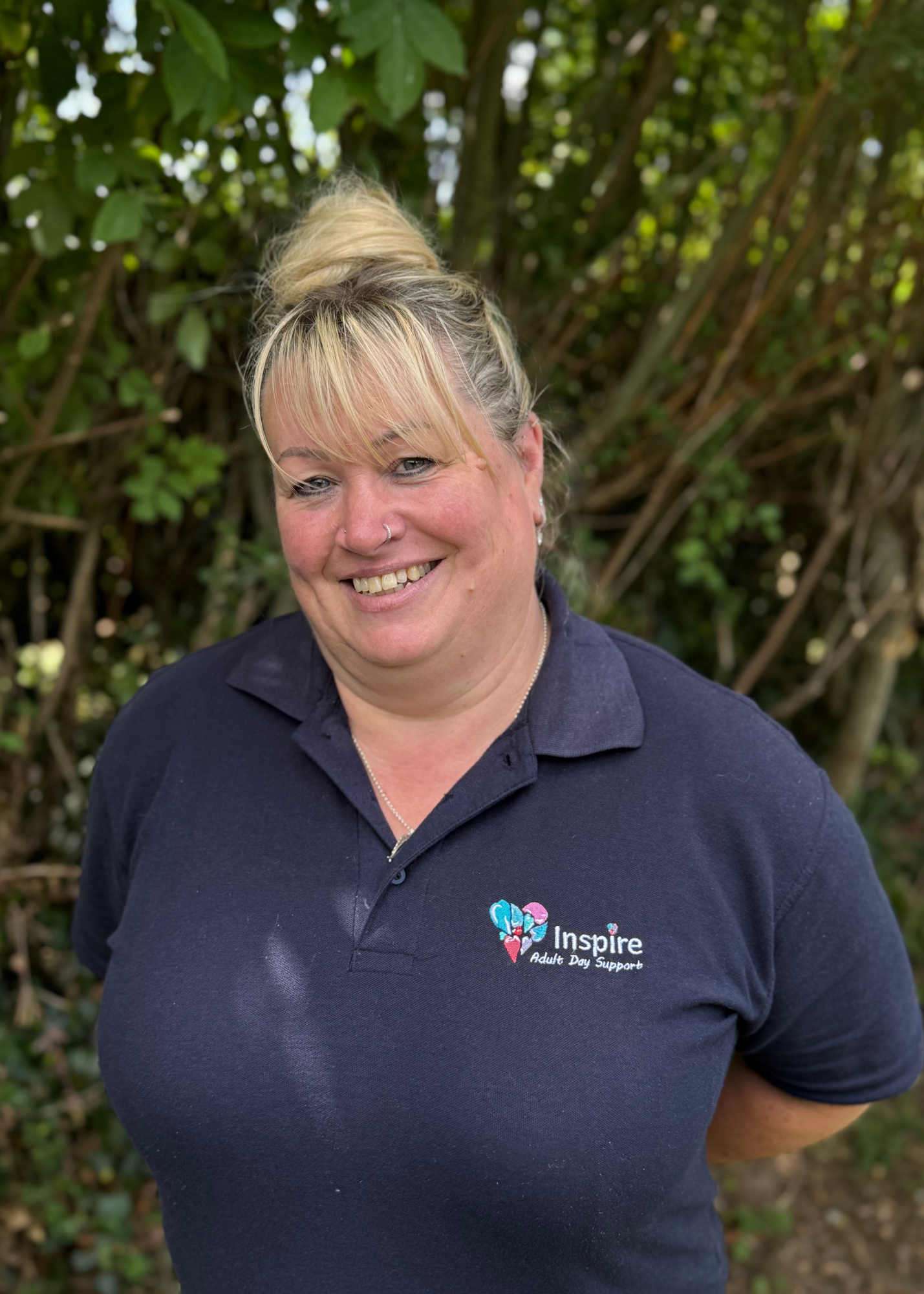 A woman outdoors wearing a blue polo shirt with an 'Inspire Adult Day Support' logo, smiling with closed eyes, background of green foliage.