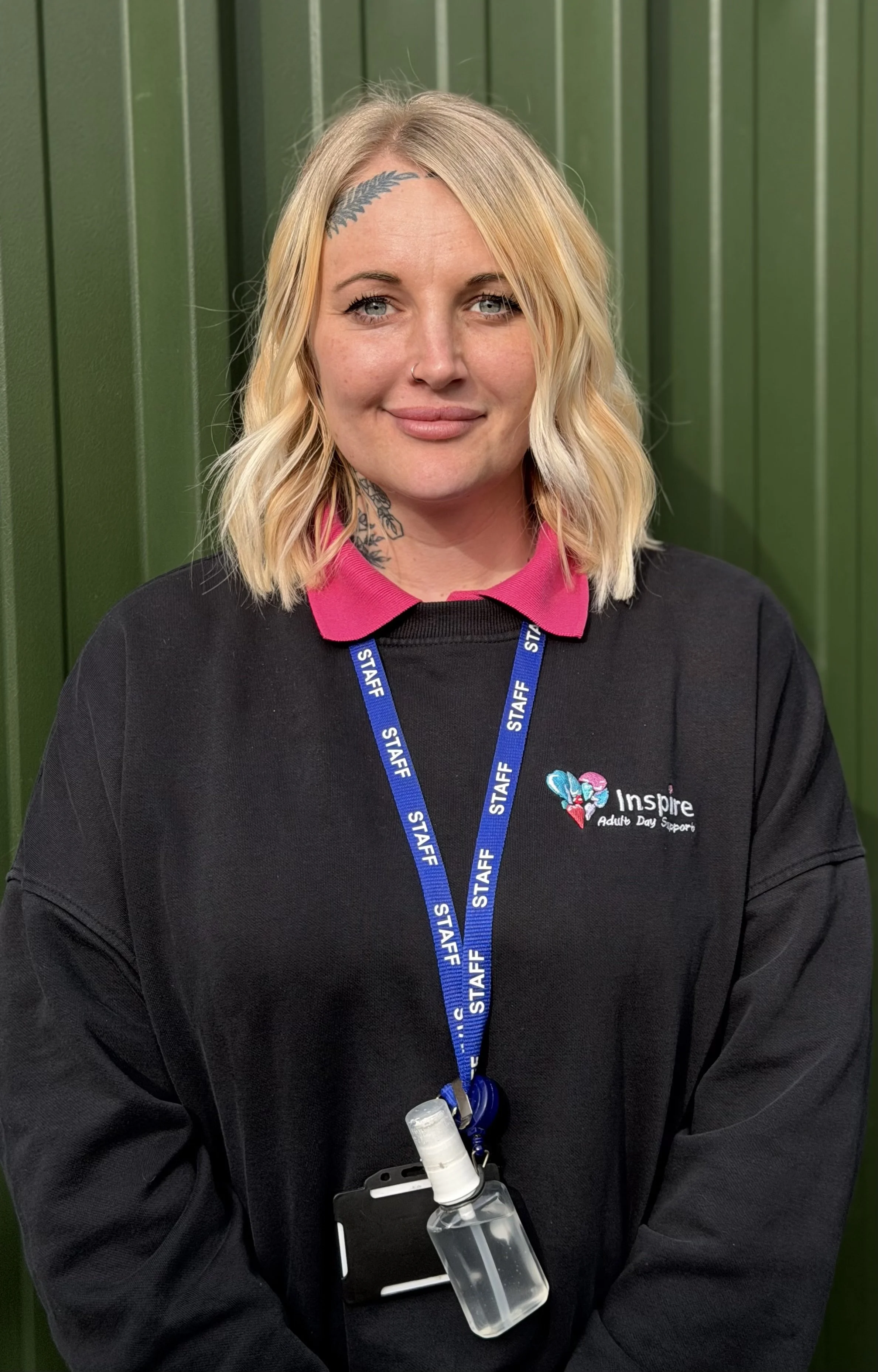 A woman with platinum blonde hair, tattoos on her neck and forehead, wearing a black polo shirt with a logo that says Inspire Adult Day Support, and a pink lanyard with white puzzle piece symbols, standing outdoors in front of green foliage.