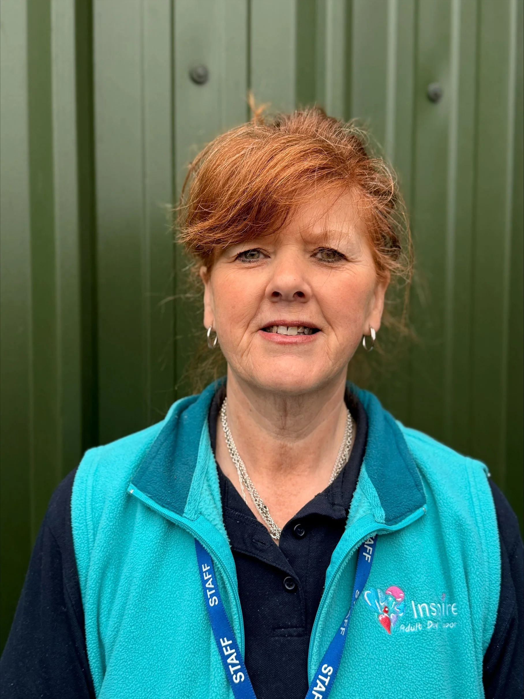 A woman with long, wavy red hair and a wide smile, wearing a navy blue polo shirt with an Inspire Adult Day Support logo, standing outdoors in front of green foliage. She has a blue lanyard with the word 'STAFF' around her neck.