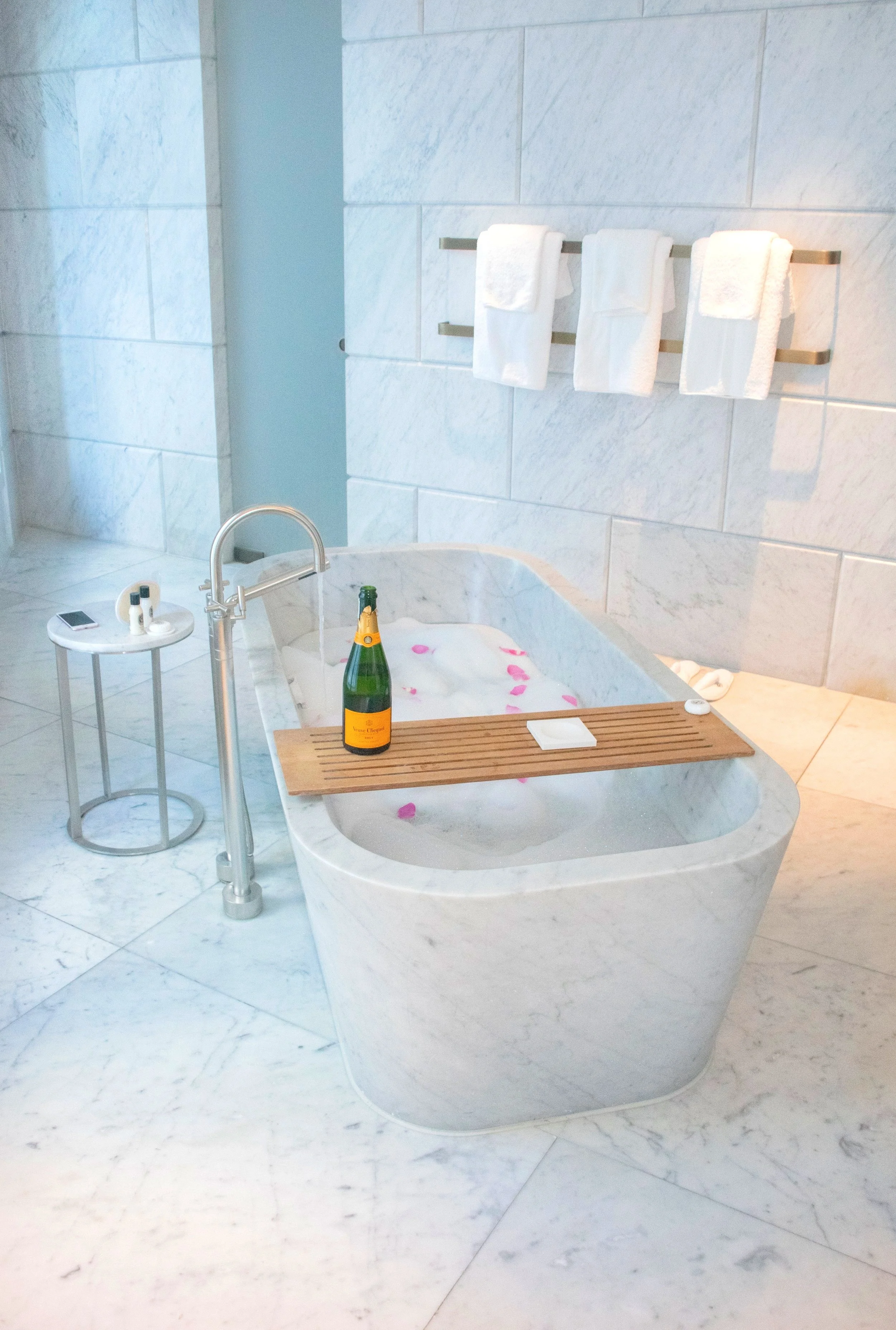 A luxurious marble bathtub with pink rose petals, a bottle of champagne, and a wooden bath tray. There are three white towels hanging on a metal towel rack on the wall behind the bathtub. A small round table beside the tub holds toiletries and a smar